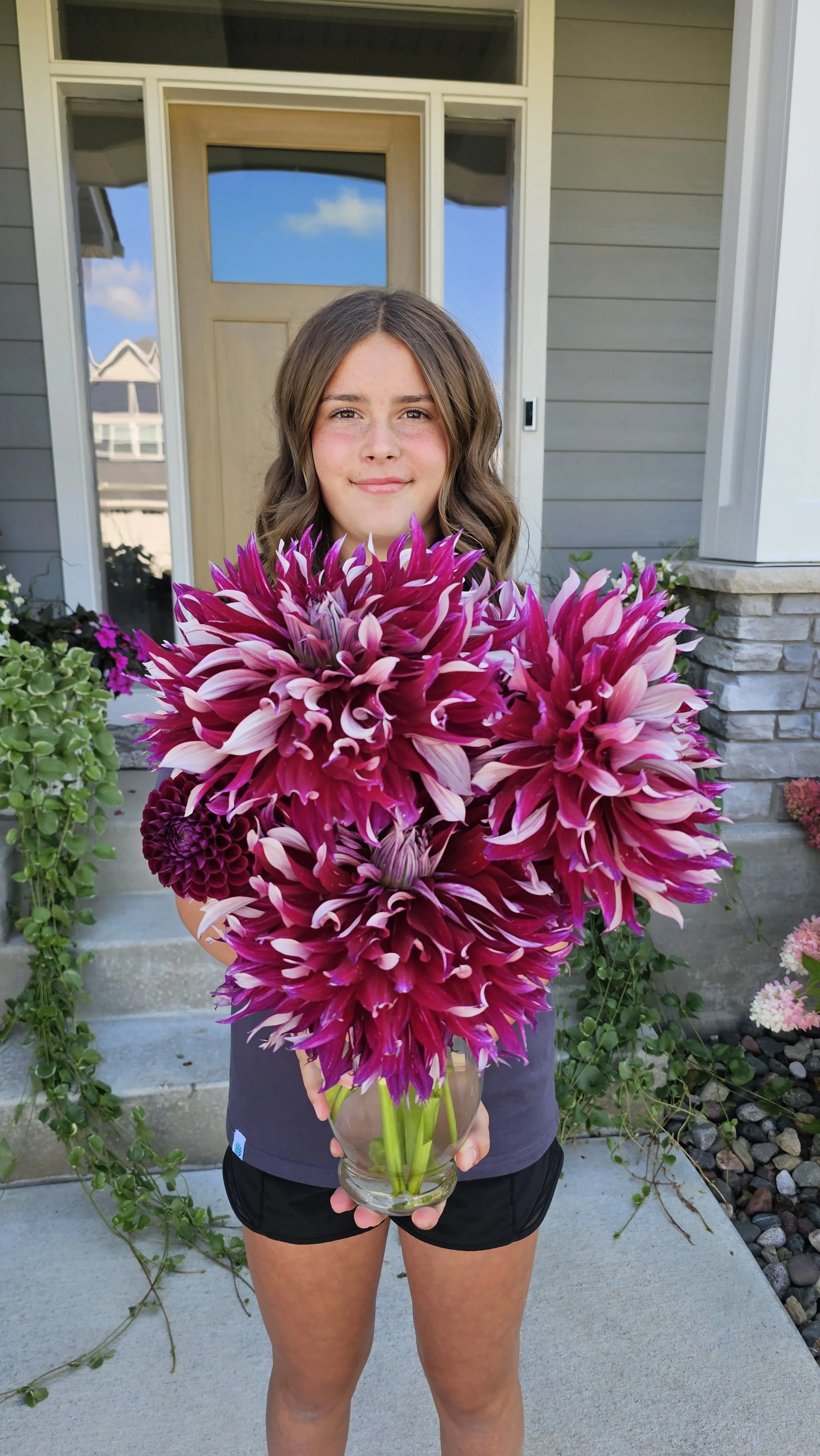A girl standing on a porch holding a large bouquet of purple and pink dahlias in a glass vase, with a house entrance behind her.