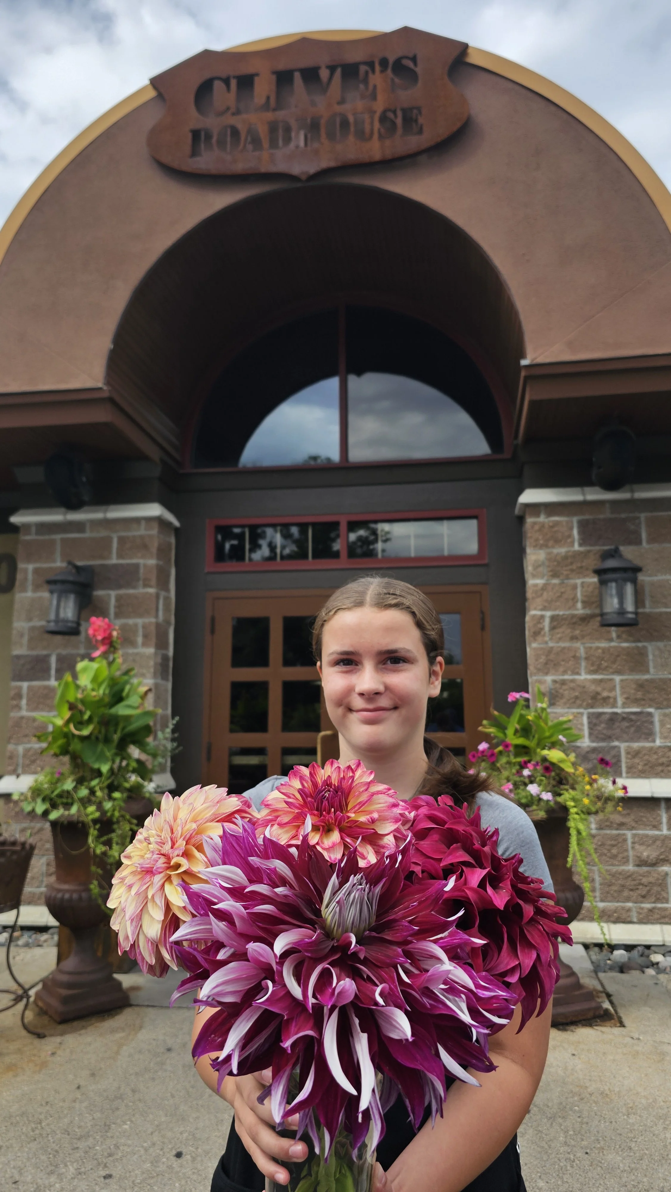 Young girl smiling holding a large bouquet of pink and purple dahlias outside in front of a building with a sign that reads "Clive's Roadhouse" and potted plants nearby.