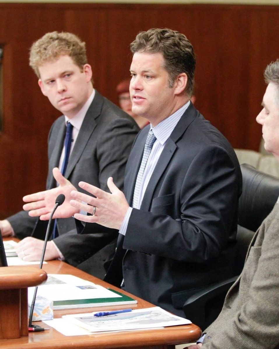 A man in a suit speaking during a formal meeting, with two other men in suits listening attentively.