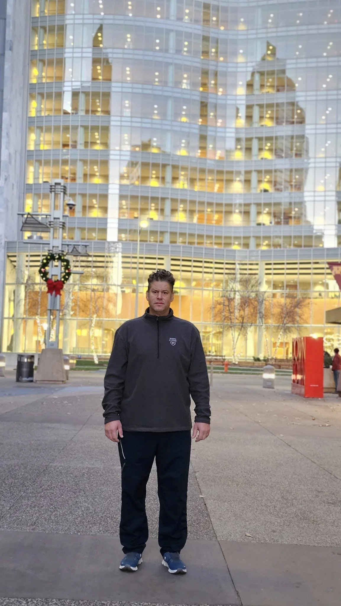 A man in black athletic clothing standing outdoors in front of a modern glass building decorated with holiday wreaths and ribbons, during the evening.
