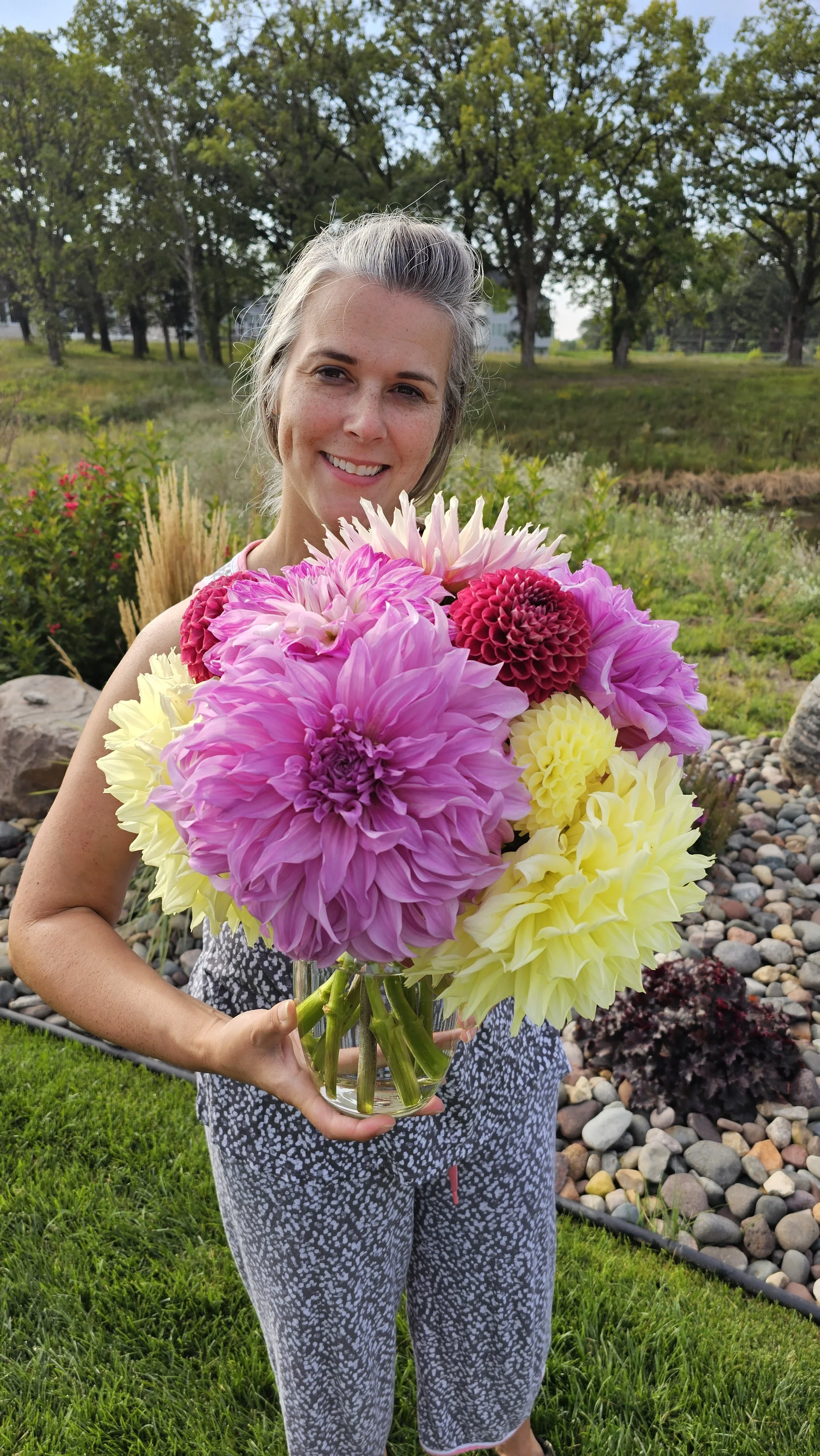 Woman holding a large colorful bouquet of dahlias outdoors in a garden with trees and rocks.