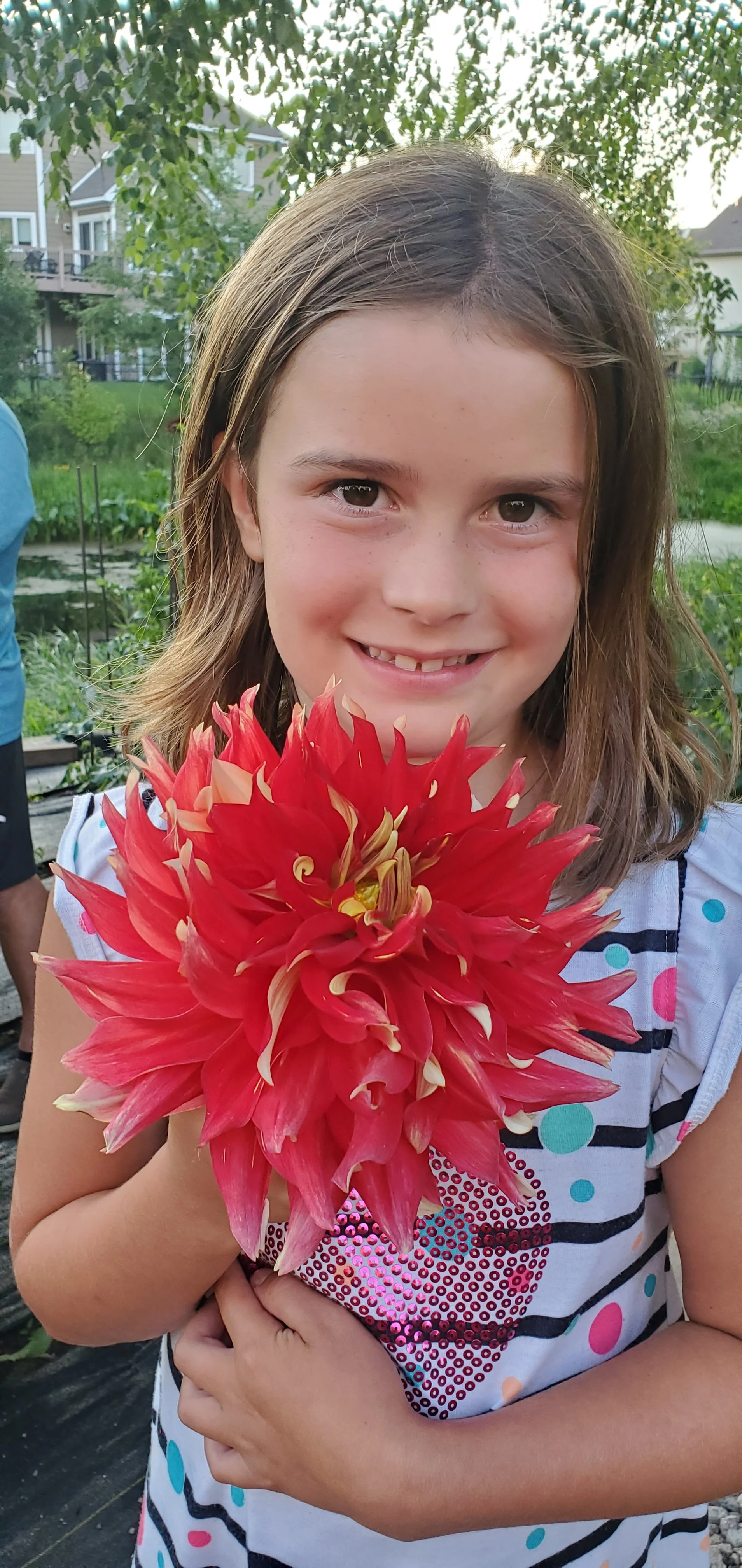 A young girl smiling and holding a large red dahlia flower outdoors in a garden.