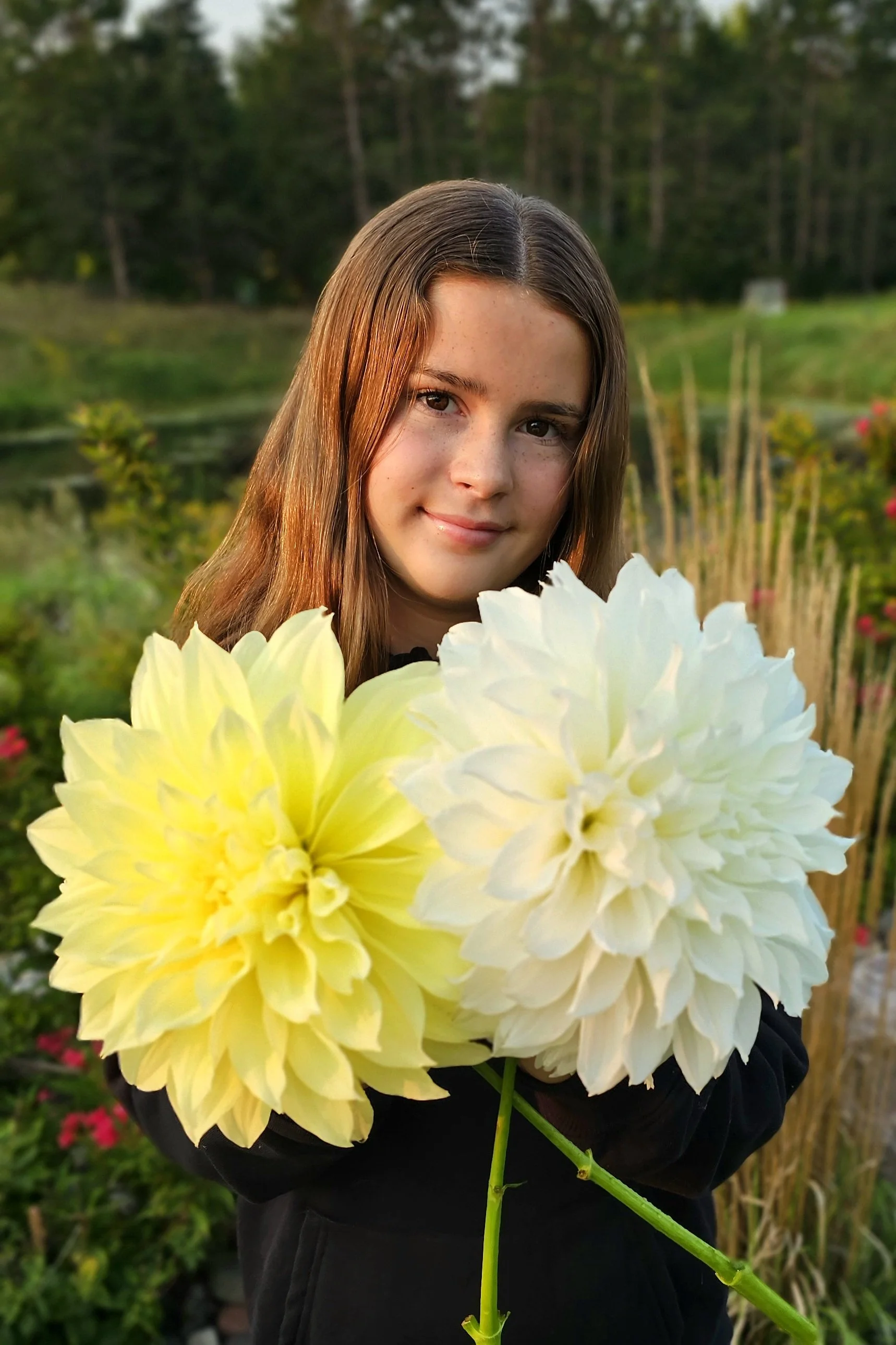 A young girl with long brown hair holding large white and yellow flowers outdoors with a background of greenery and trees.
