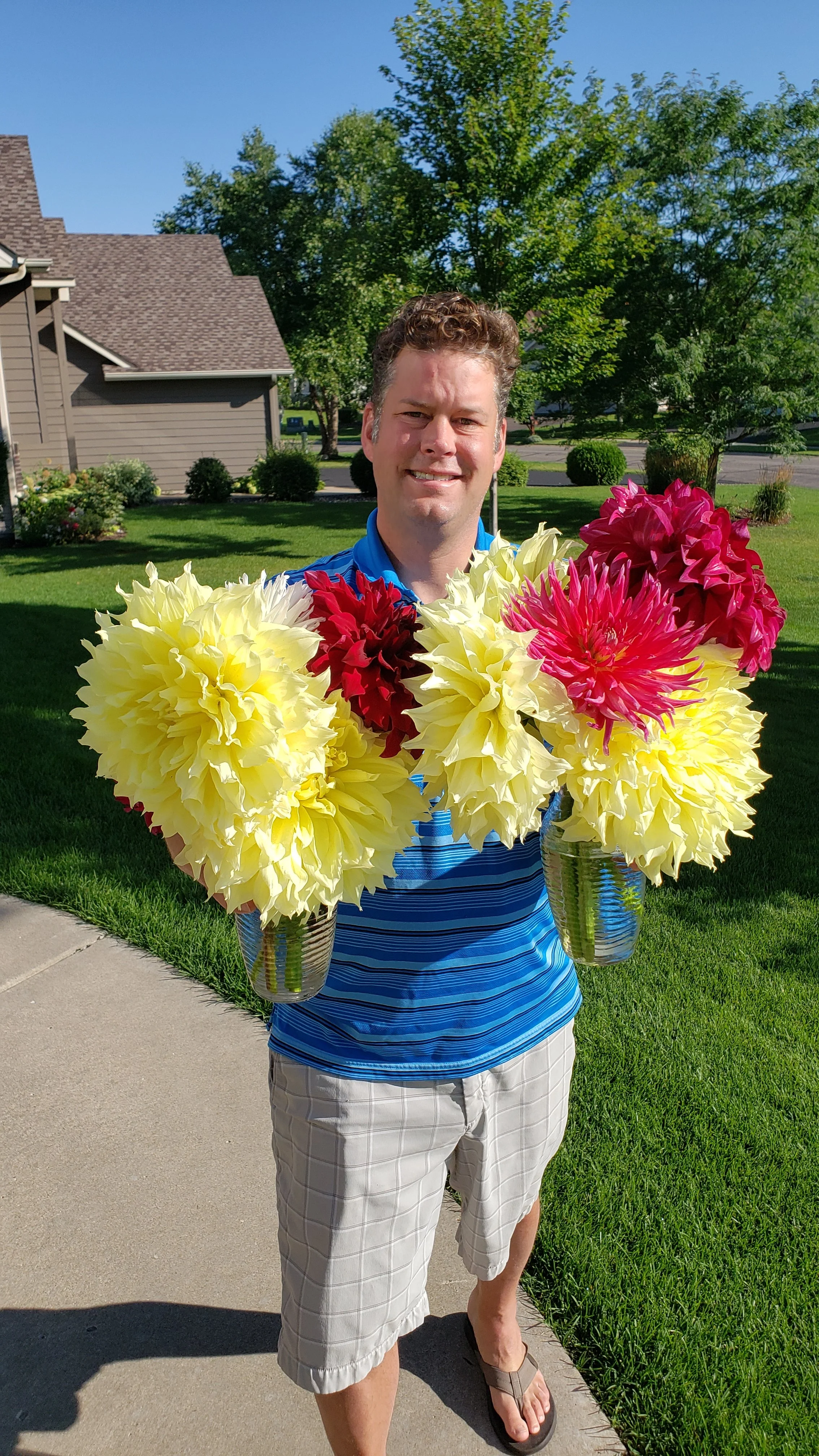 A man standing on a sidewalk in a suburban neighborhood holding two vases with large, colorful flowers in each hand, smiling at the camera.