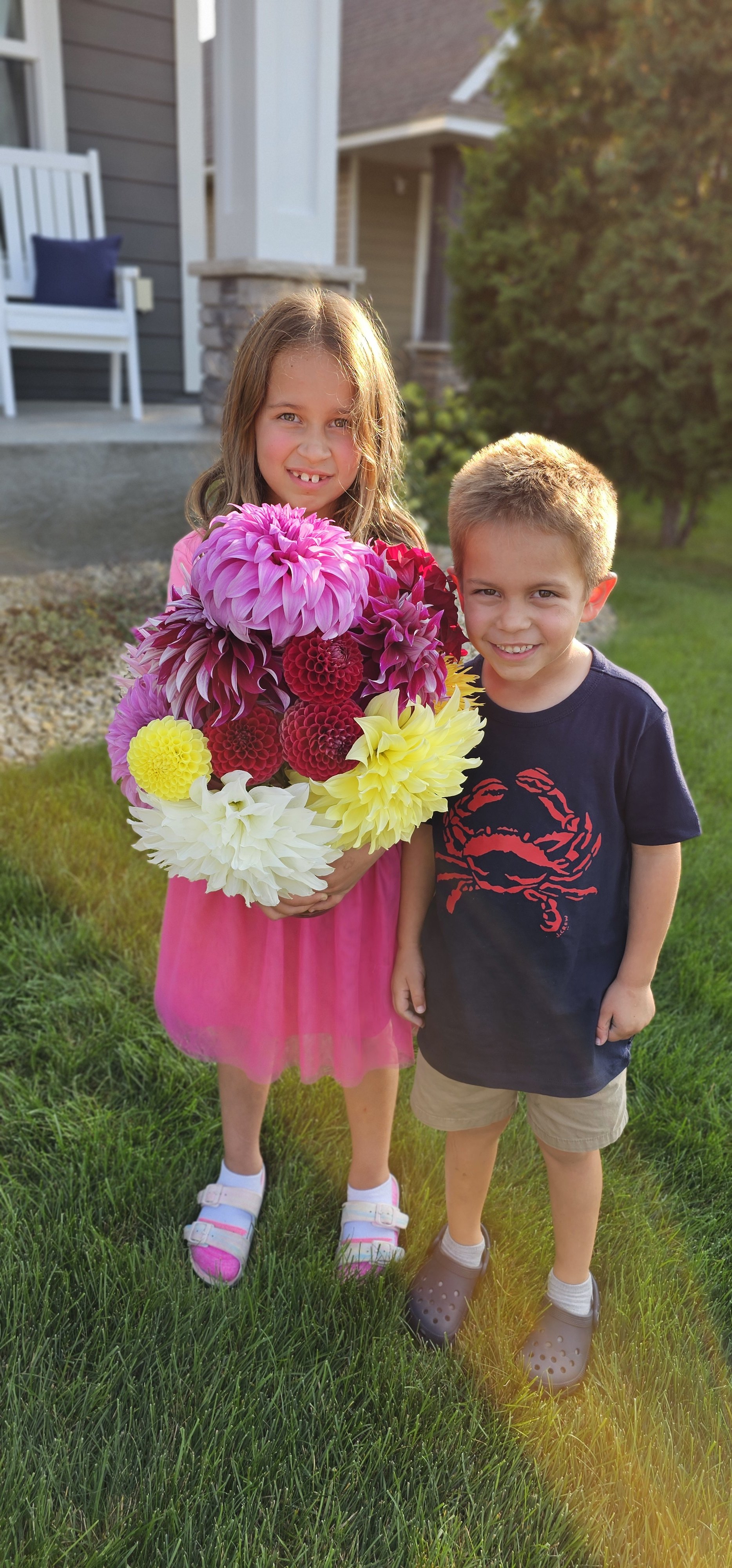 A young girl and boy standing on grass in front of a house, holding a large bouquet of colorful flowers, with the girl wearing a pink dress and sneakers and the boy wearing a navy t-shirt with a crab graphic and khaki shorts.