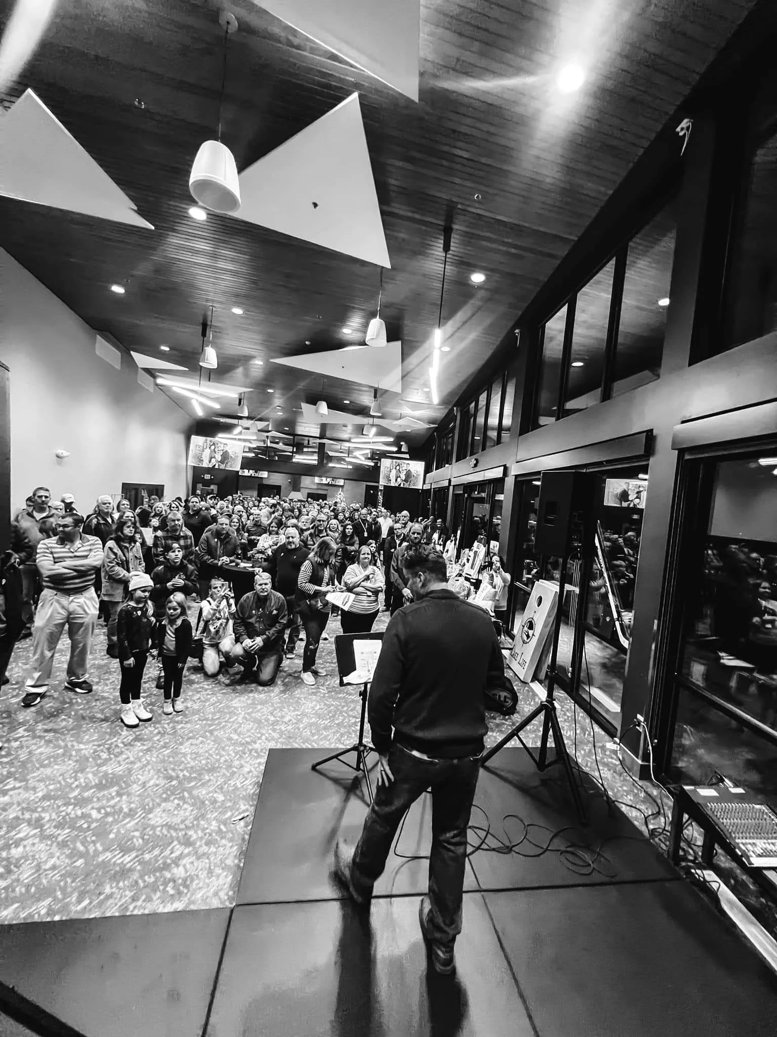 A crowded indoor event space with a speaker performing on stage in front of a large audience. The room has wooden ceilings and modern lighting fixtures, with some screens and posters visible in the background.