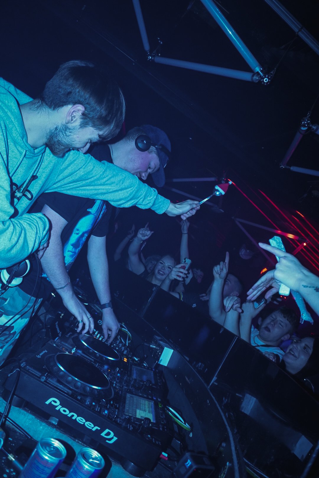 Two DJs perform at a nightclub with a crowd dancing and celebrating, illuminated by blue and red lights.