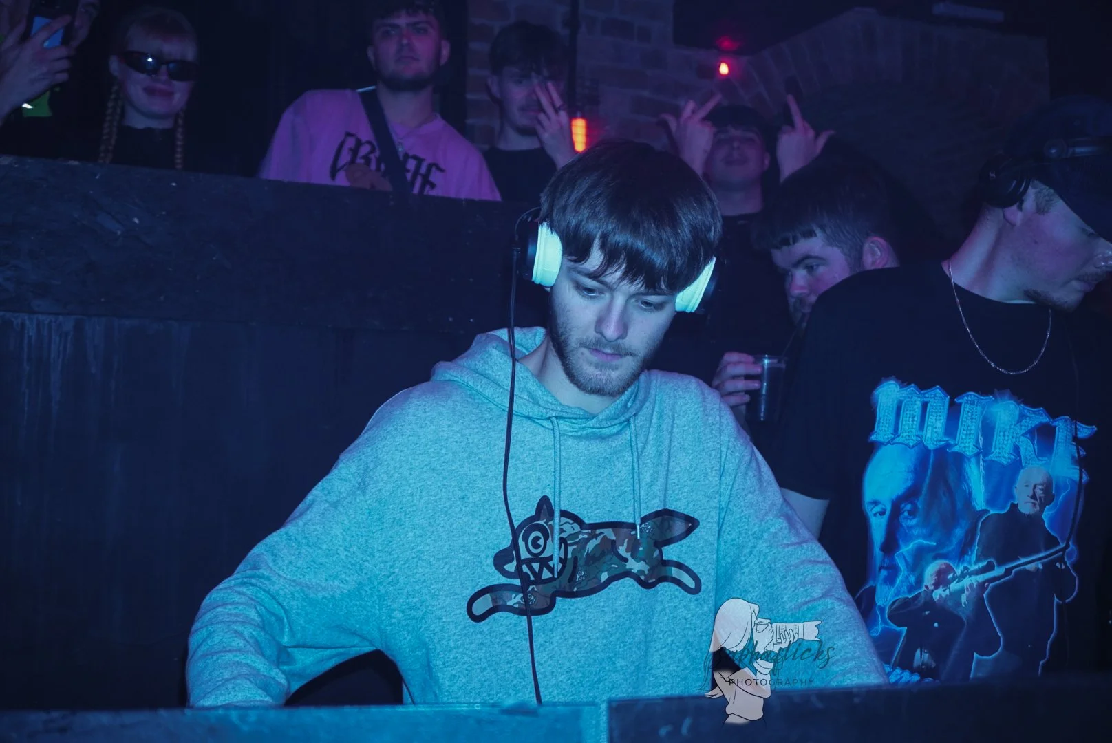 A young man with brown hair and a beard, wearing a gray hoodie with a cartoonish dog or wolf graphic, is DJing at a nightclub with headphones on. Several people are in the background, some making hand gestures and holding drinks, in a dimly lit envir