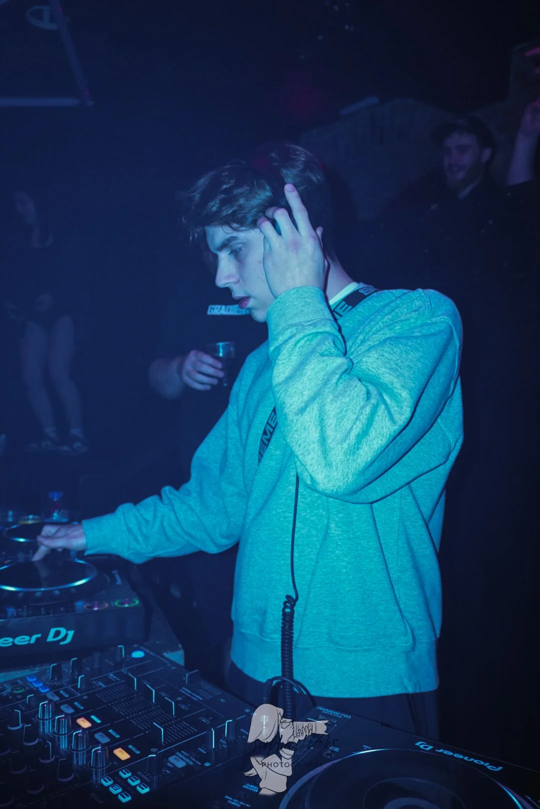 Young male DJ wearing a gray hoodie, headphones, and a race car driver lanyard, performing at a nightclub with DJ equipment and a dark, smoky atmosphere.