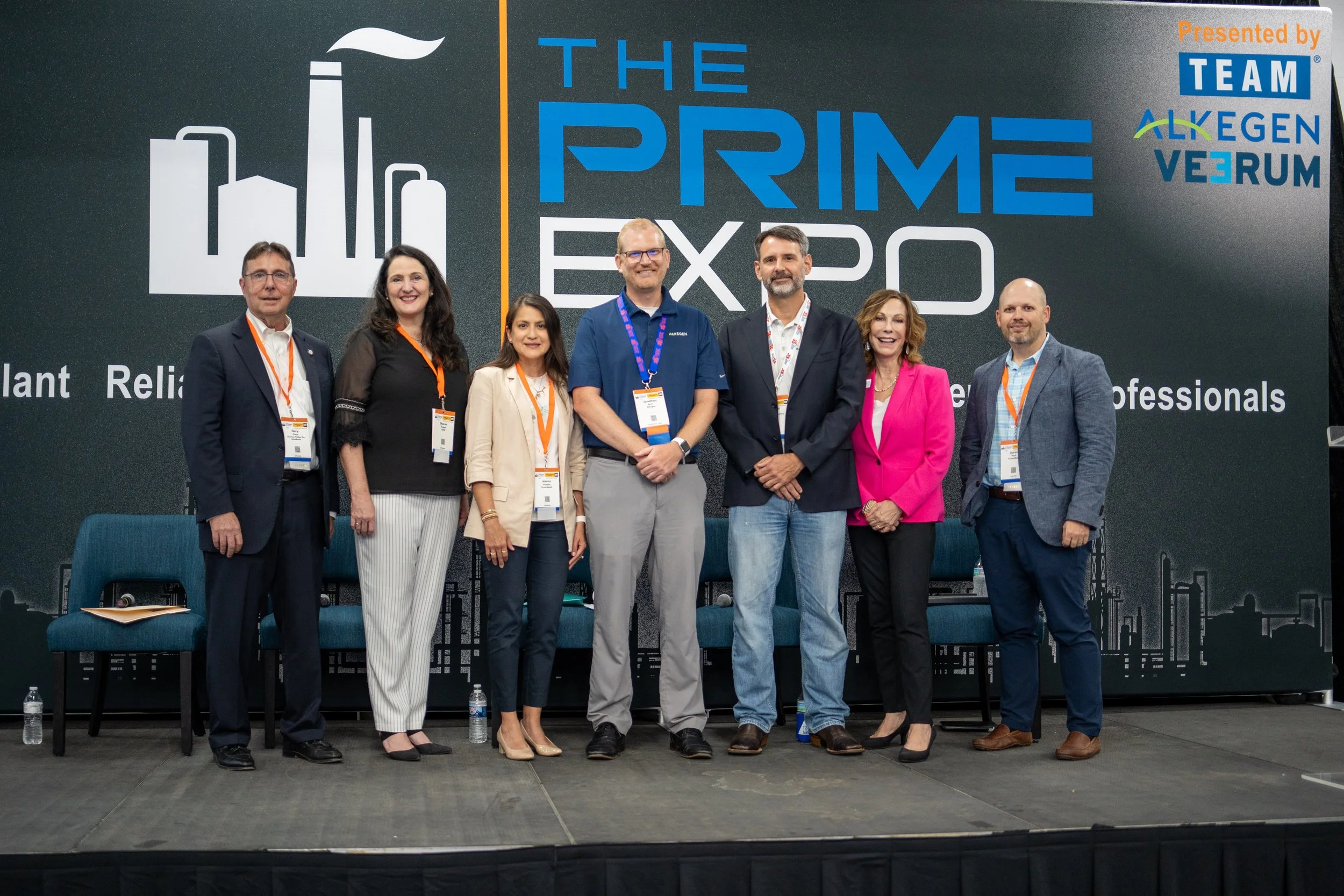 Group of eight professionals standing on stage at The Prime Expo, with a large backdrop displaying event branding and logos, including Team AlkeGEN VeRUM.