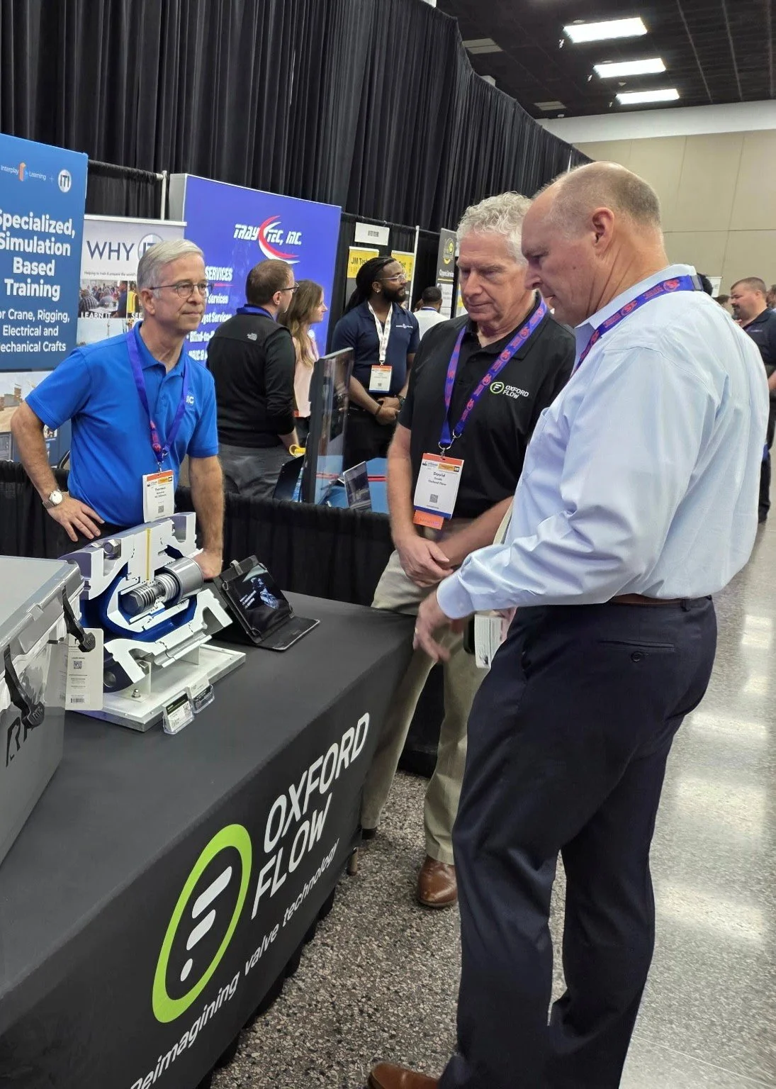 A trade show booth for Oxford Flow featuring a display of mechanical components, with three men engaged in conversation, one of them explaining the products. There are banners in the background advertising specialized training and services, and the b
