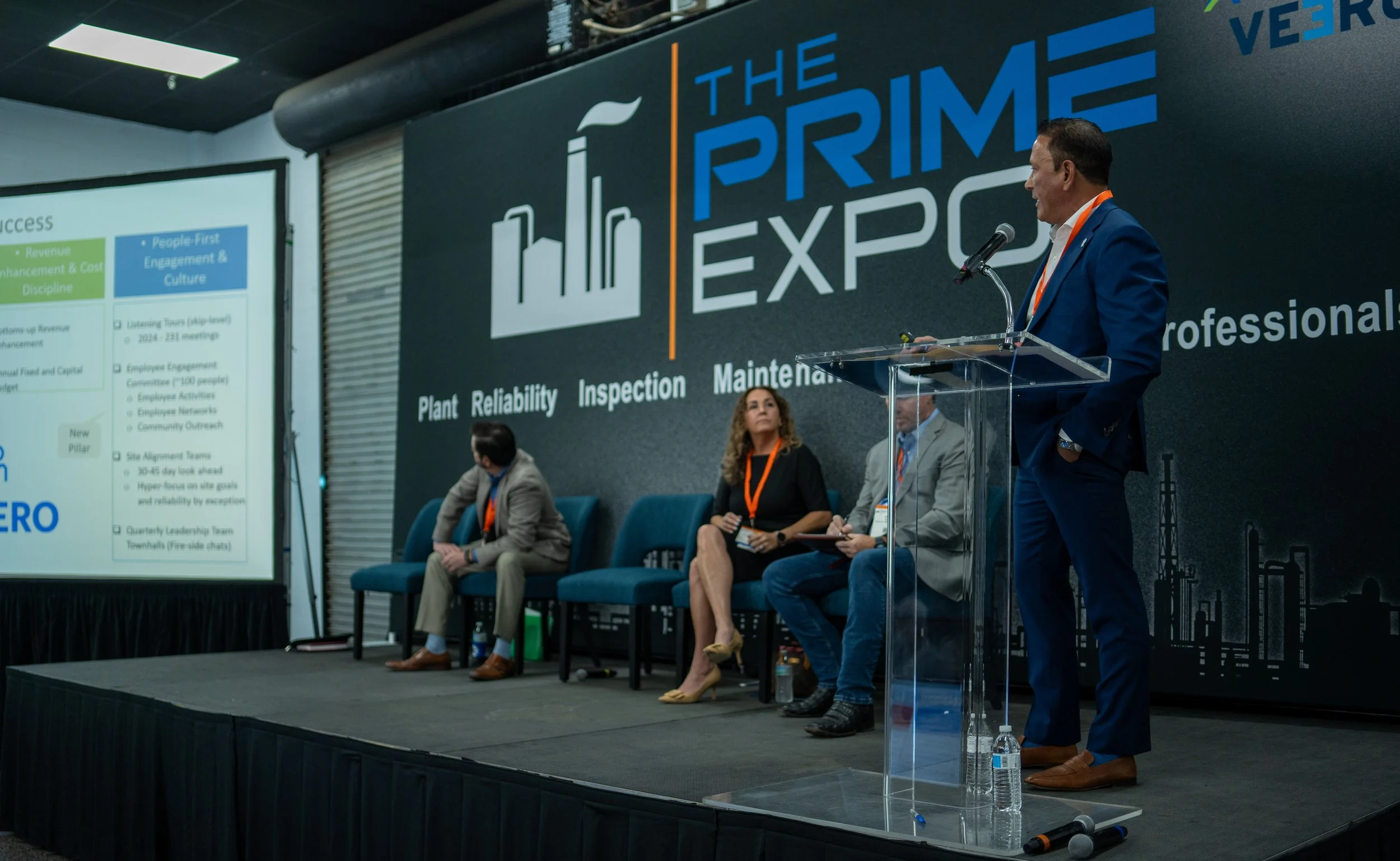 A man in a blue suit speaks at a podium during a conference, with four seated panelists behind him on a stage. The background features a large sign reading 'The Prime Expo' with themes 'Plant Reliability, Inspection, Maintenance.' A presentation slid