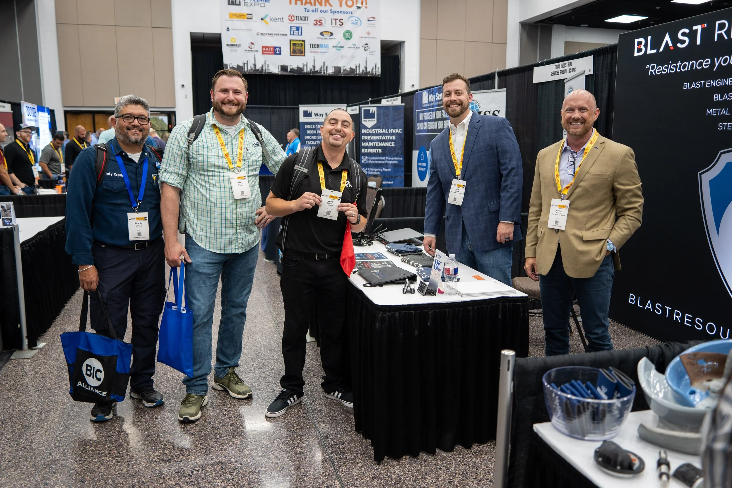 Group of five men standing at a trade show booth, smiling at the camera, with banners and booths in the background.
