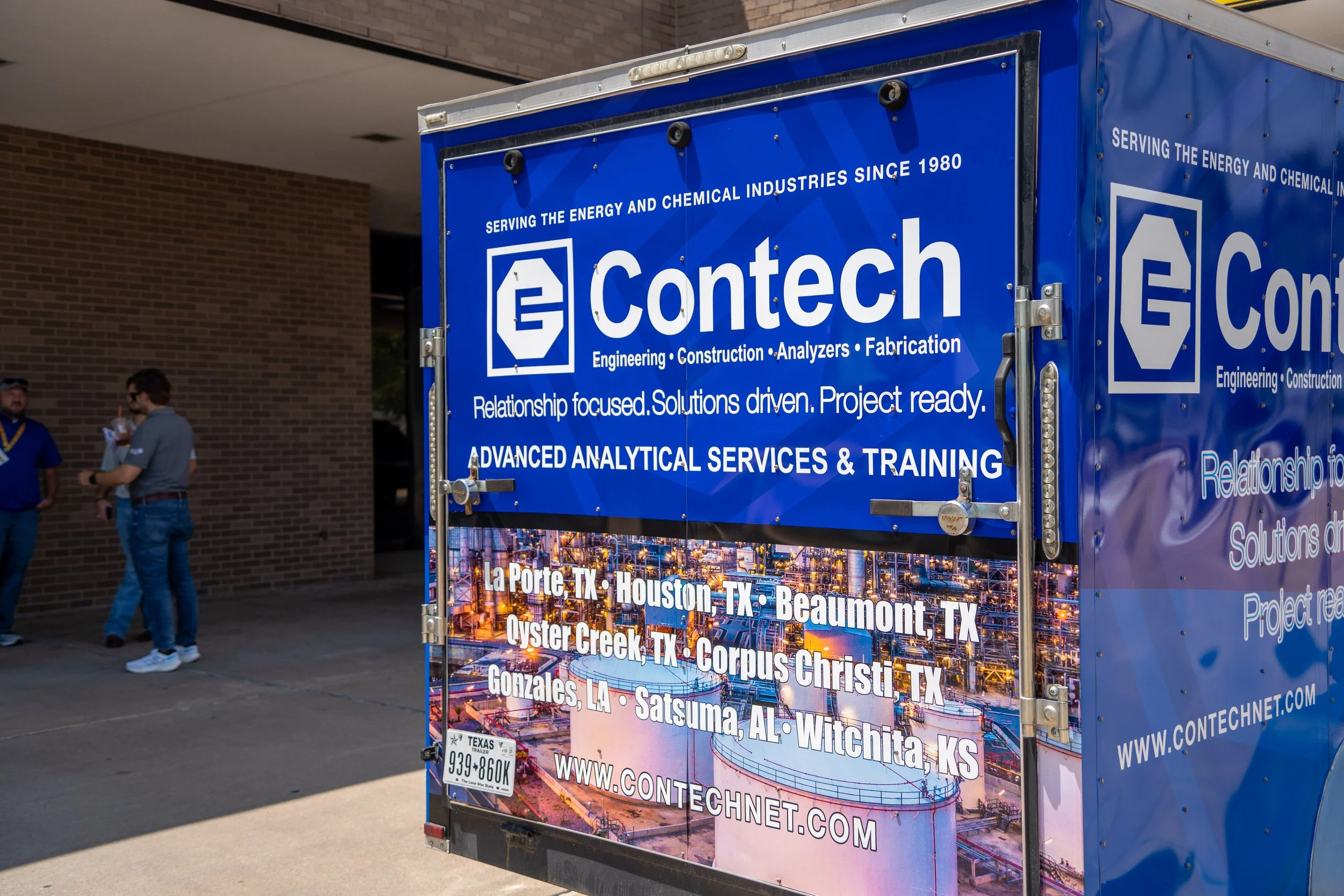 Blue Contech truck with company information, list of Texas cities served, and a website address, parked outside a brick building with people standing nearby.