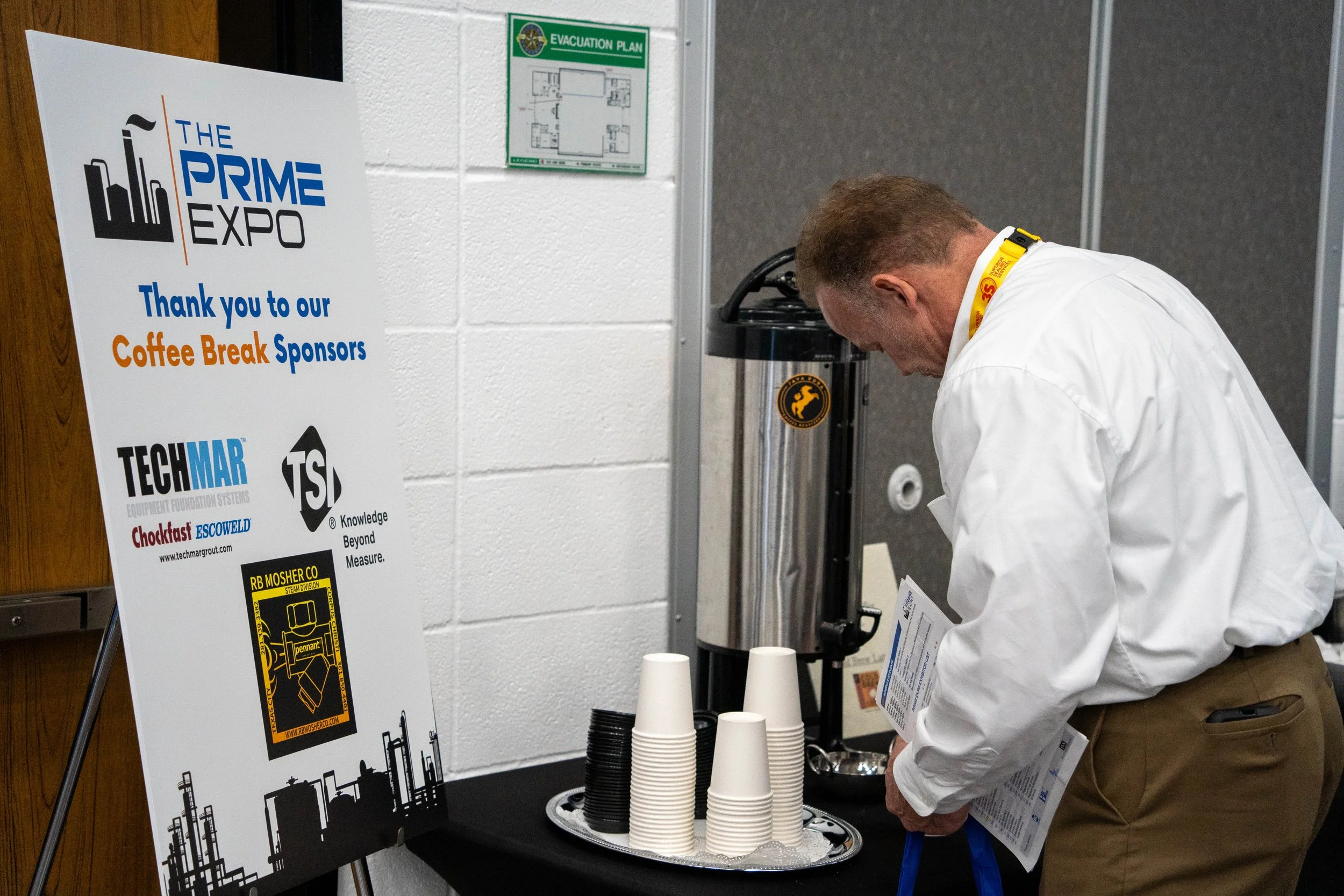 A man in a white shirt and brown pants is getting coffee from a coffee station, which includes cups, a thermos, and a sign thanking sponsors at The Prime Expo.