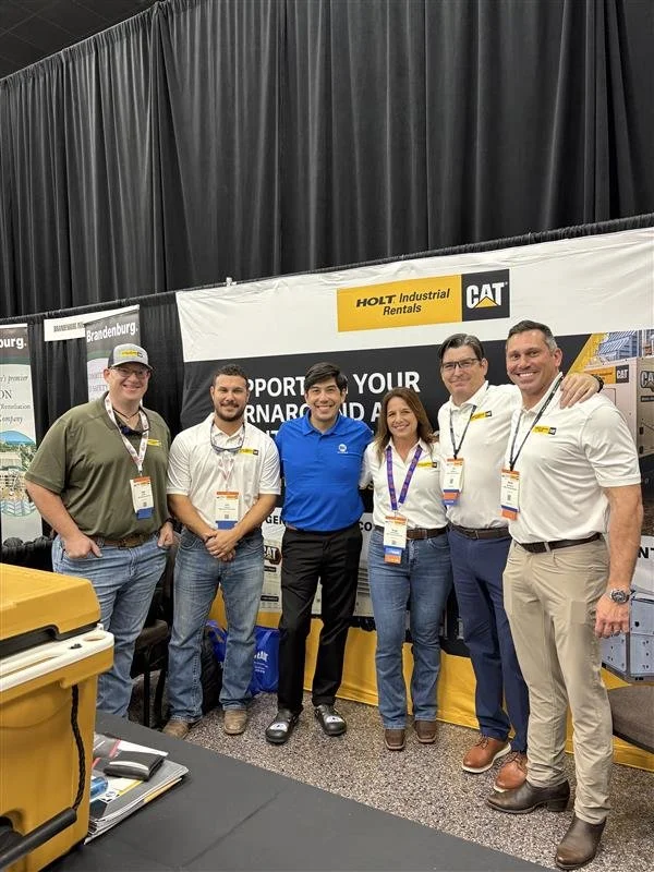 Group of six people standing in front of a booth at an event, with banners displaying 'HOLT Industrial Rentals' and 'CAT'.