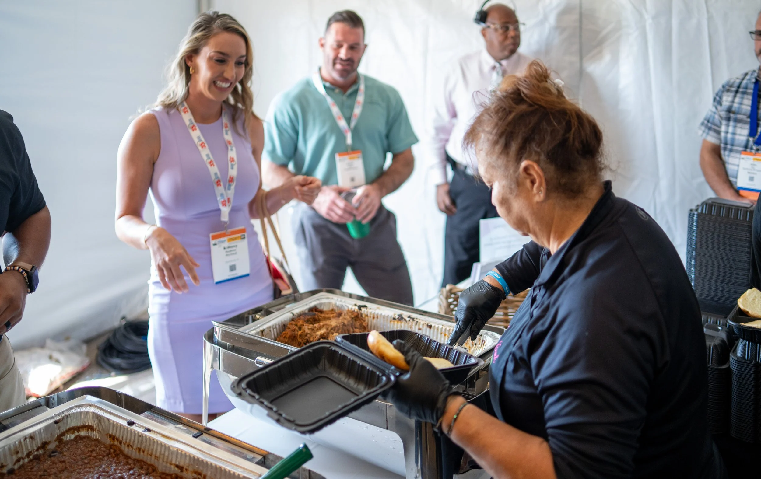 A woman in a lavender dress is serving food from a buffet tray at a food event, while several people in the background watch and wait.