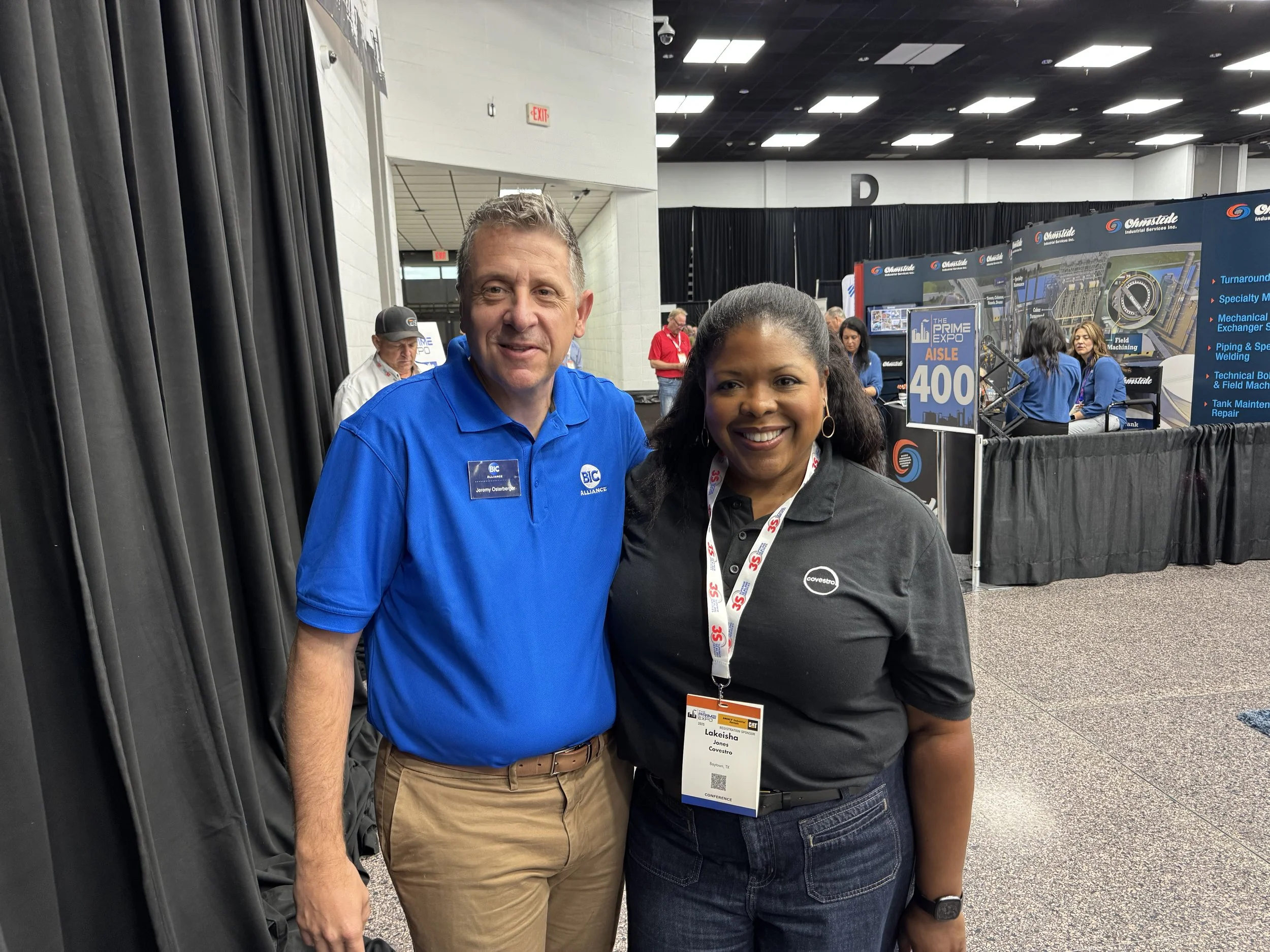Two professionals smiling at a trade show, the man on the left wearing a blue polo shirt with a name tag and the woman on the right wearing a black polo shirt with a conference badge, in front of exhibition booths with people and banners in the backg
