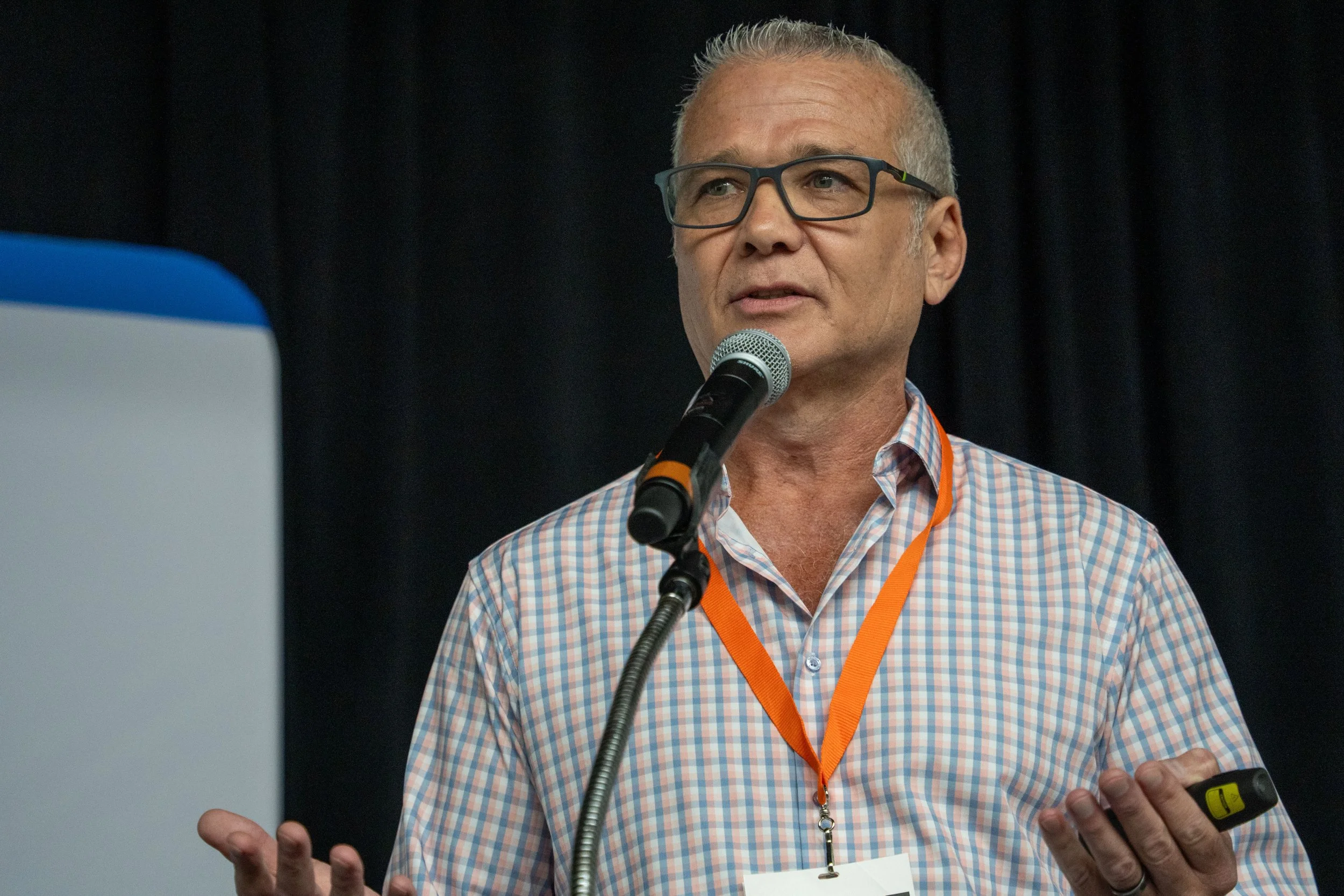 A middle-aged man with glasses and short gray hair speaking into a microphone while holding a remote control. He's wearing a checkered shirt and an orange lanyard with an ID badge.