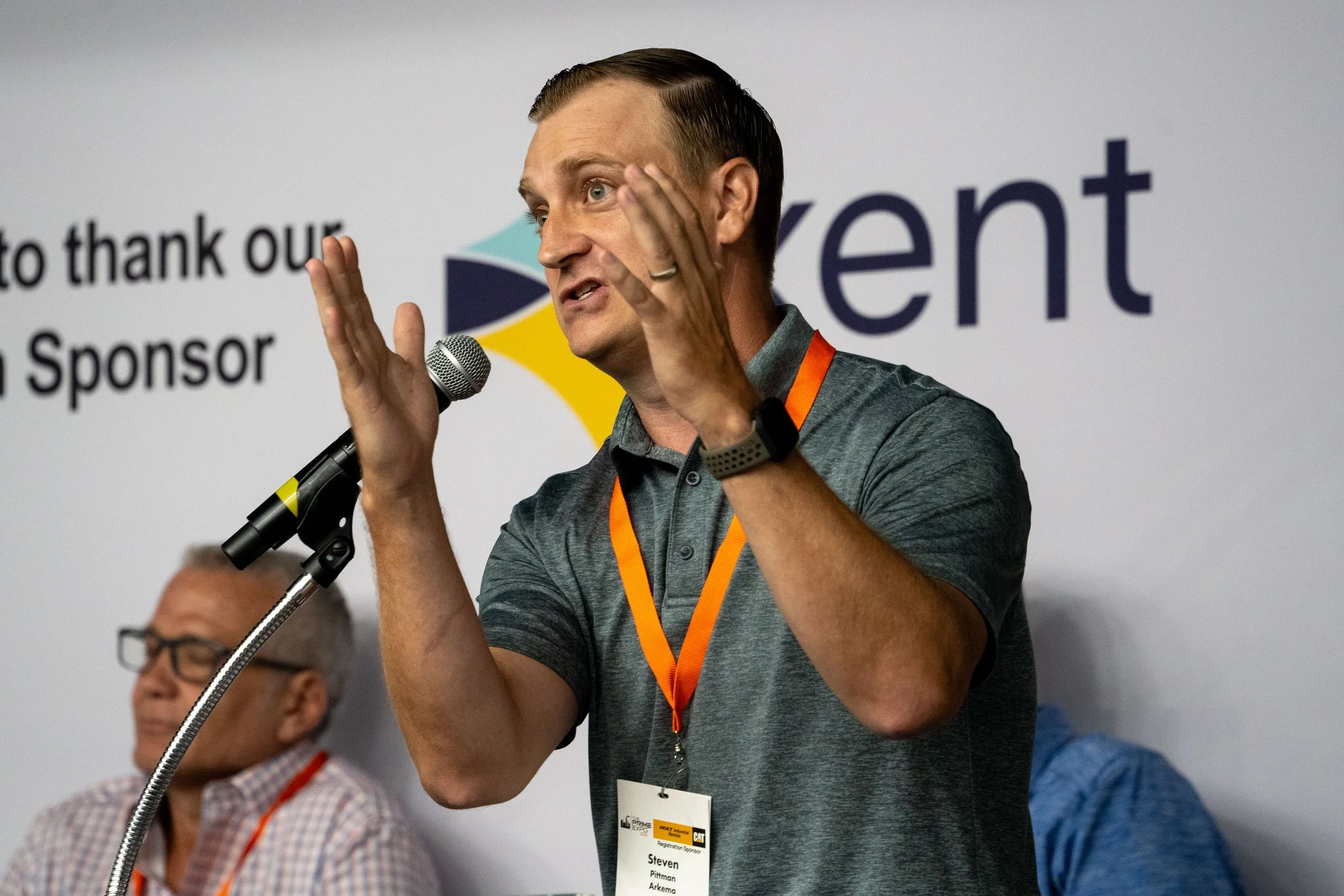 A man in a gray polo shirt with a name tag speaking into a microphone at a conference, gesturing with his hands, with a large logo and text in the background.