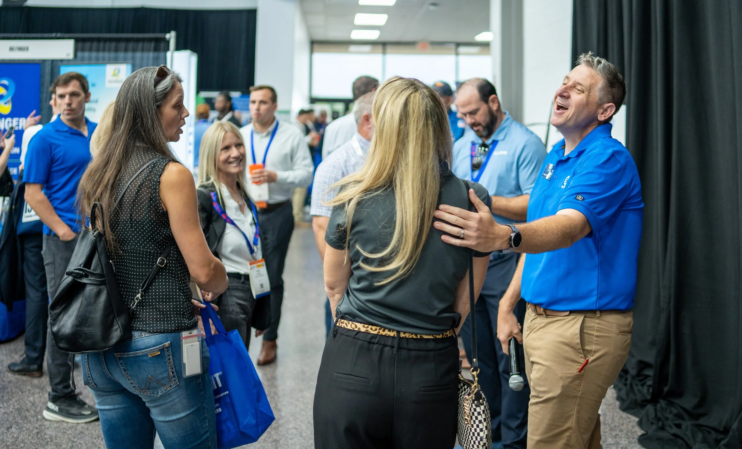 Group of people at a conference or networking event, engaged in conversation, with some smiling and others listening, in an indoor exhibition space.