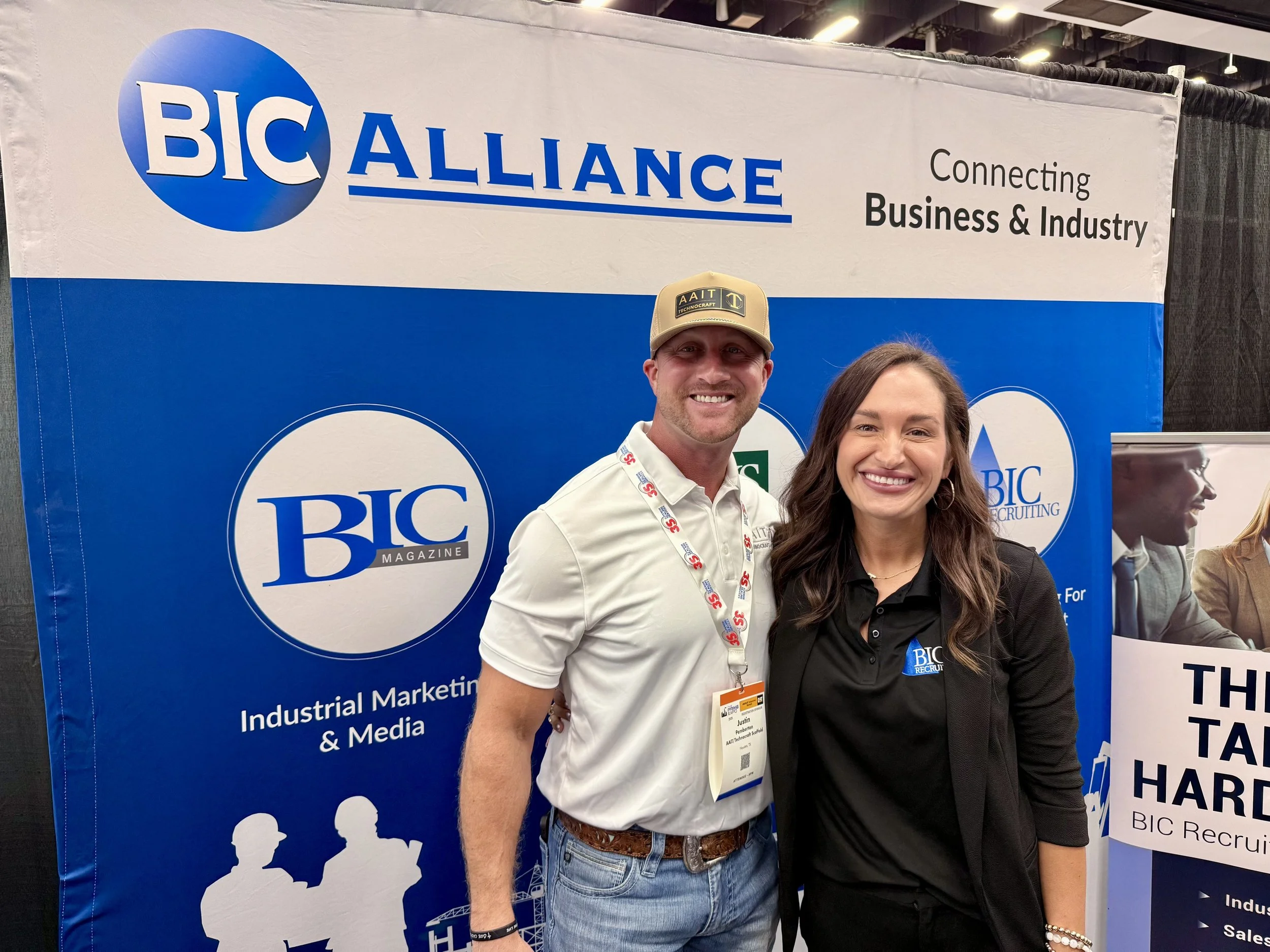 A man and woman standing together at a BIC Alliance booth during an industry event. The man wears a beige cap and white shirt, and the woman wears a black blazer and shirt. Both are smiling for the camera.