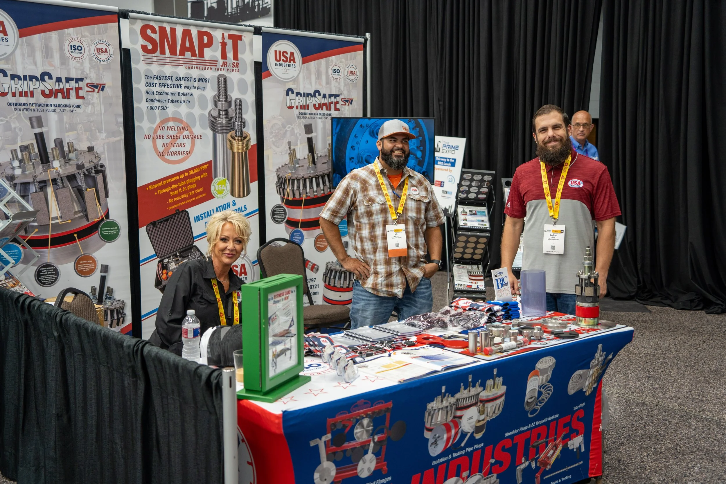 Trade show booth featuring three people standing and sitting behind a display table with industrial tools and materials. The booth has large banners with product advertisements and information. The woman is seated, and the two men are standing, smili