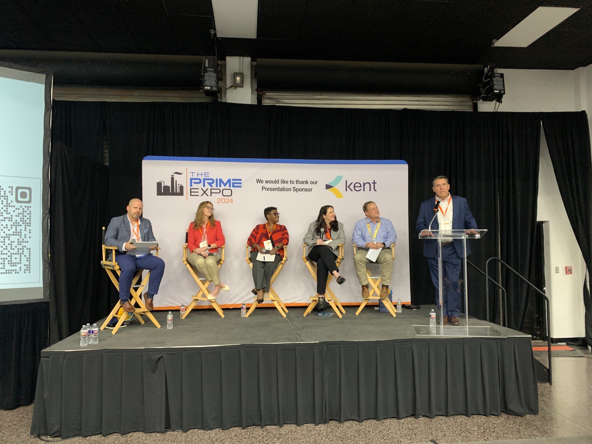 A panel discussion at The Prime Expo 2024, with five people seated on chairs on stage, one person standing at a clear podium speaking, with a backdrop displaying the event's logo and the word 'Kent'.