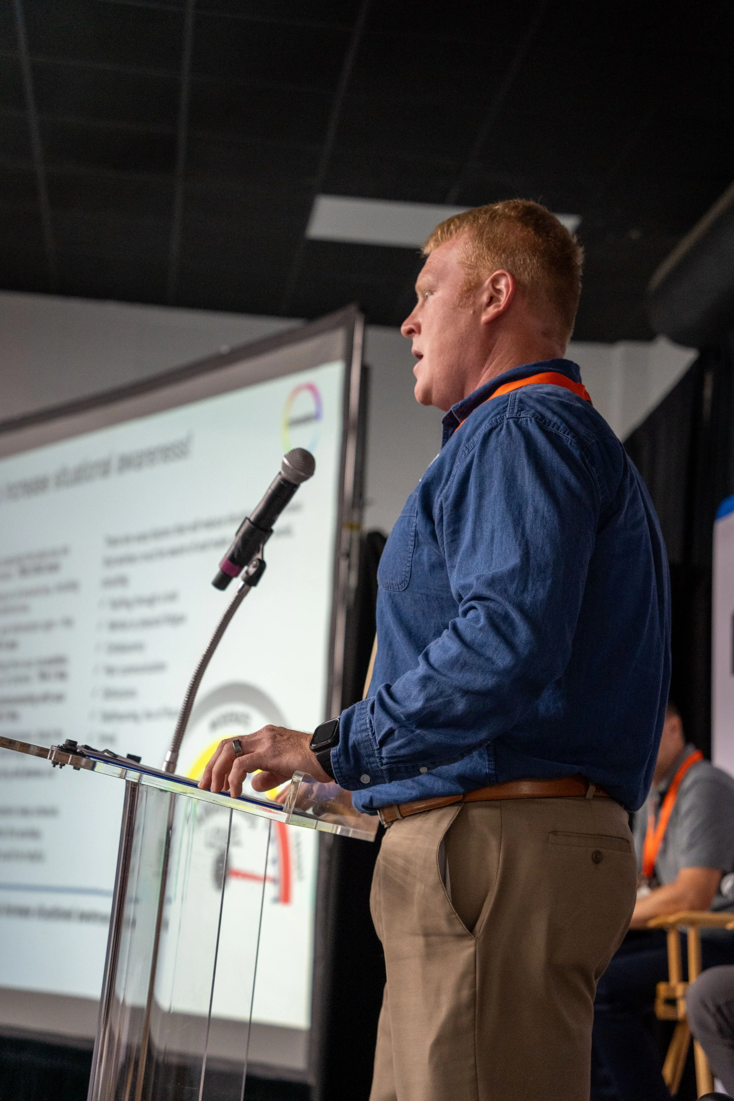 A man with red hair wearing a blue shirt and khaki pants presenting at a podium during a conference.