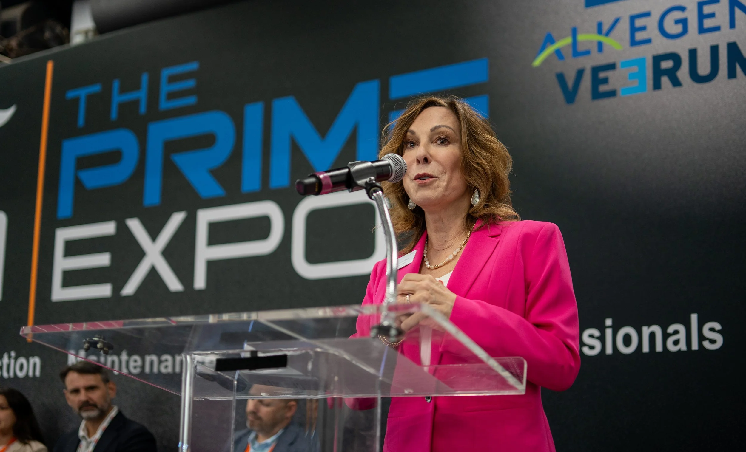 A woman with wavy reddish-brown hair wearing a pink blazer speaking at a podium with a microphone, in front of a large display board that reads 'The Prime Expo' and 'Alkegen Verum'.