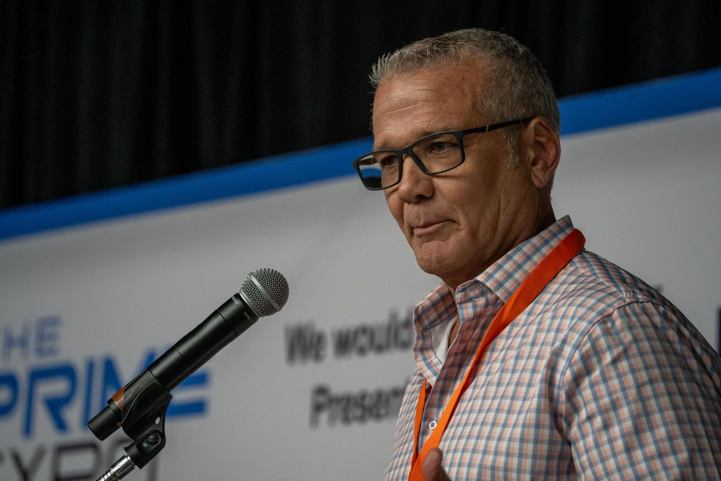 A man with short gray hair and glasses speaking into a microphone at a conference, wearing a checkered shirt and an orange lanyard.