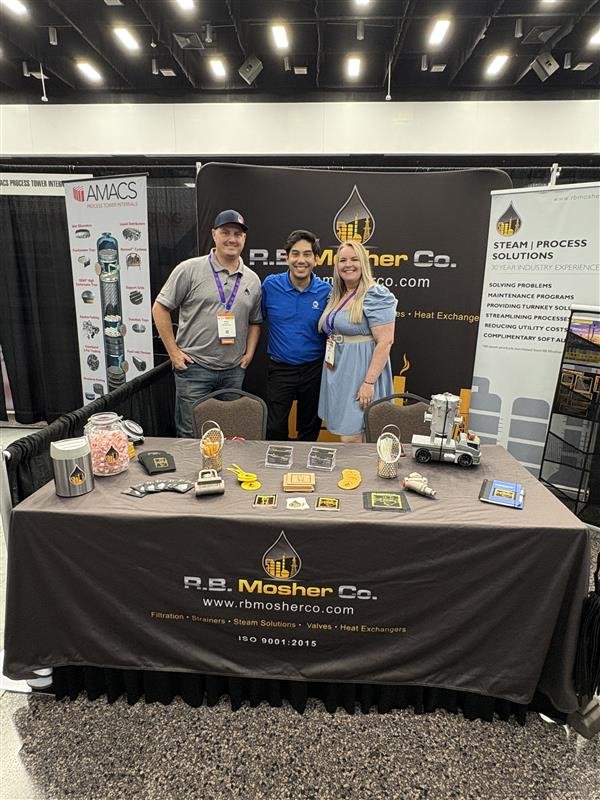 Three people standing behind a table with various industrial equipment and promotional materials at a trade show.