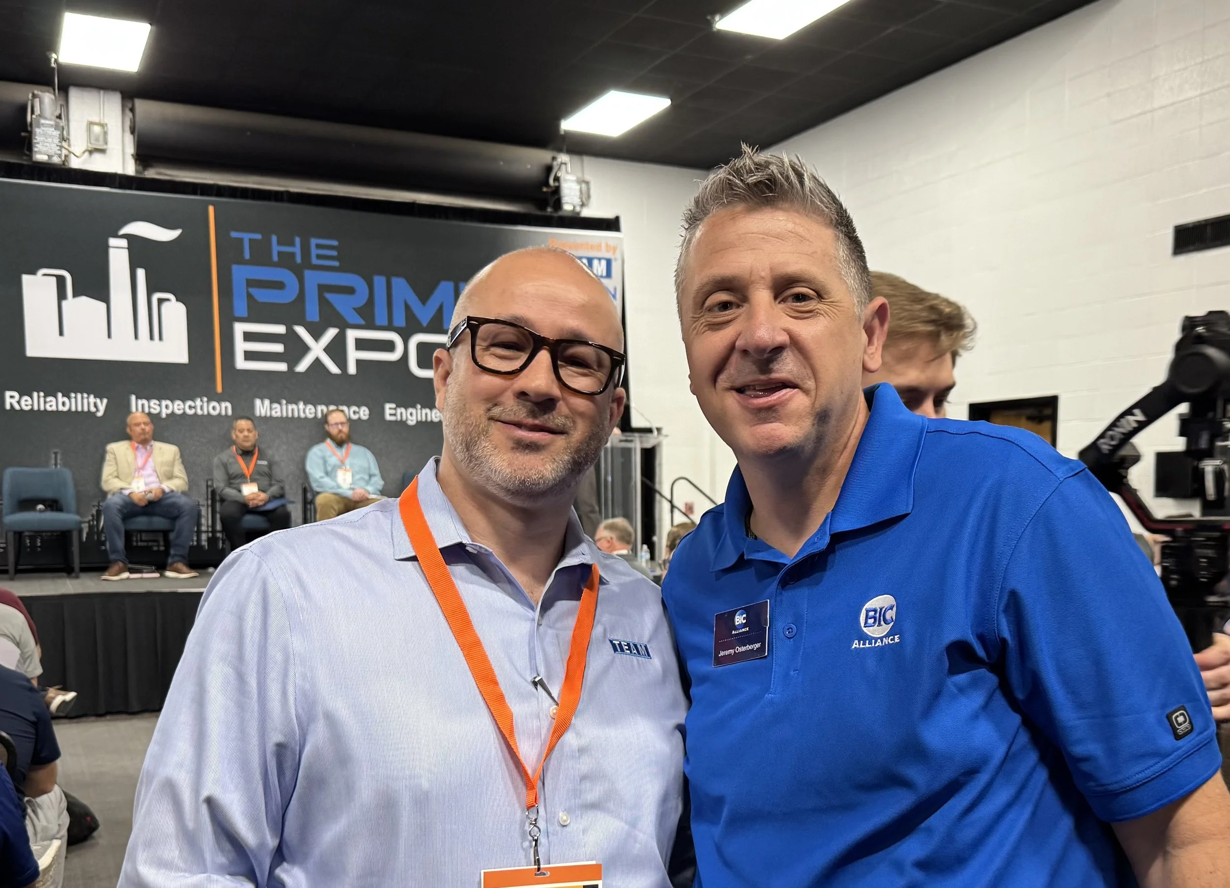 Two men smiling at the Primex Conference, with a stage in the background featuring five seated panelists and a large sign reading 'The Primex Expo'.