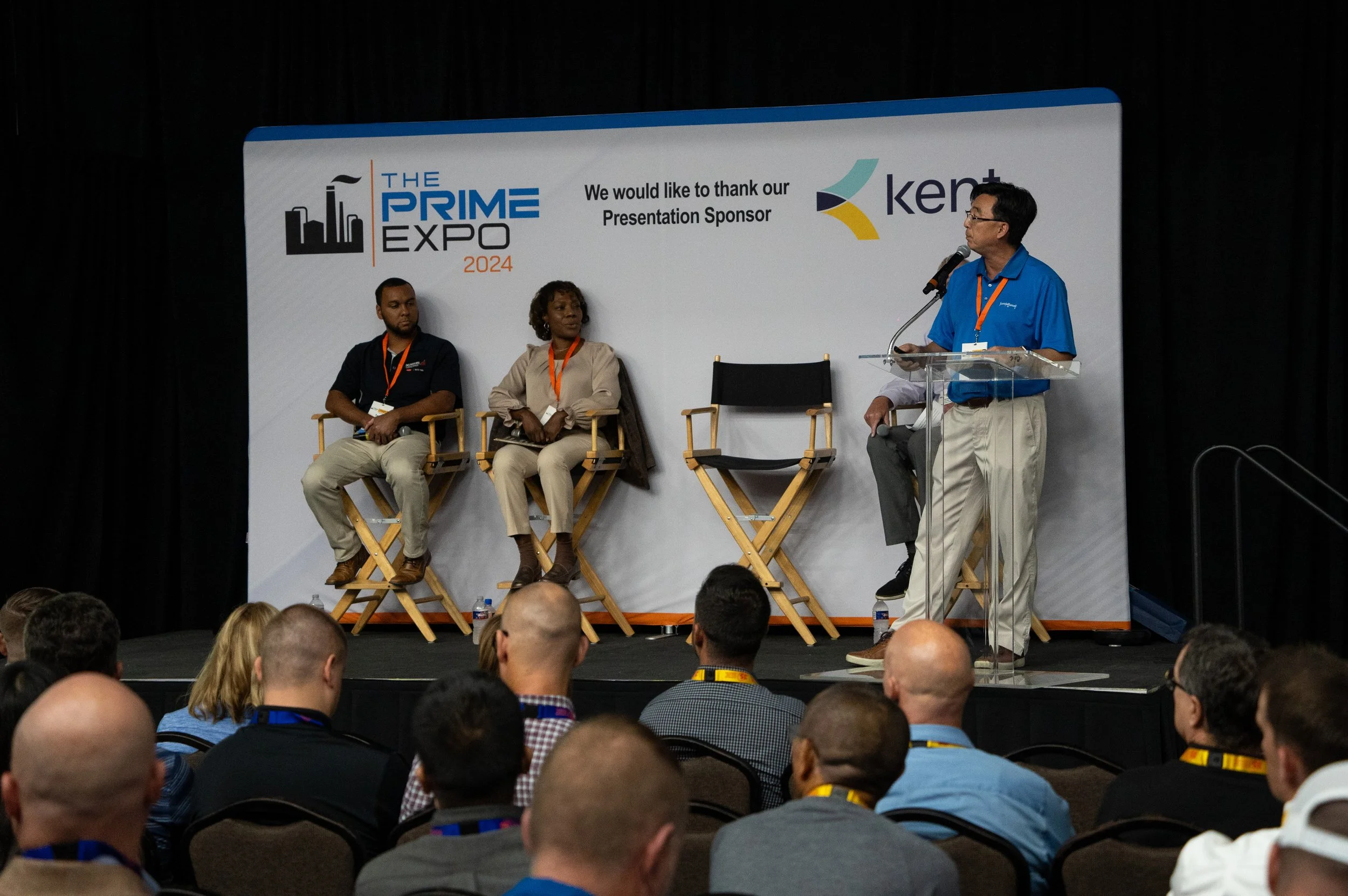 Panel discussion at The Prime Expo 2024, with four individuals on stage, one standing at a clear podium speaking into a microphone, three sitting in director's chairs, and an audience watching. The stage backdrop features logos and a thank you messag