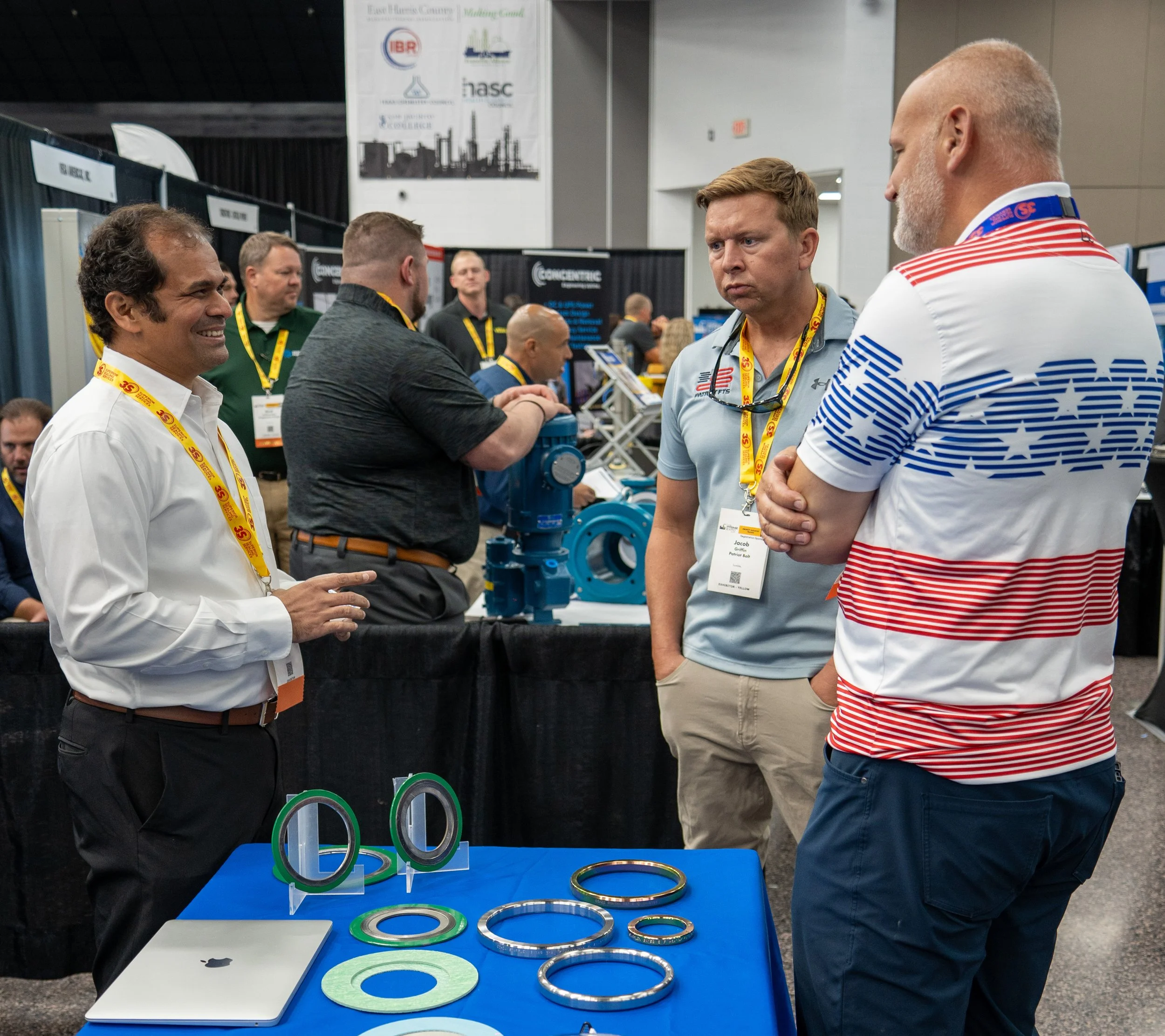 Three men engaged in conversation at a trade show, with a table displaying industrial seals and an open laptop. Other attendees and exhibition booths are visible in the background.