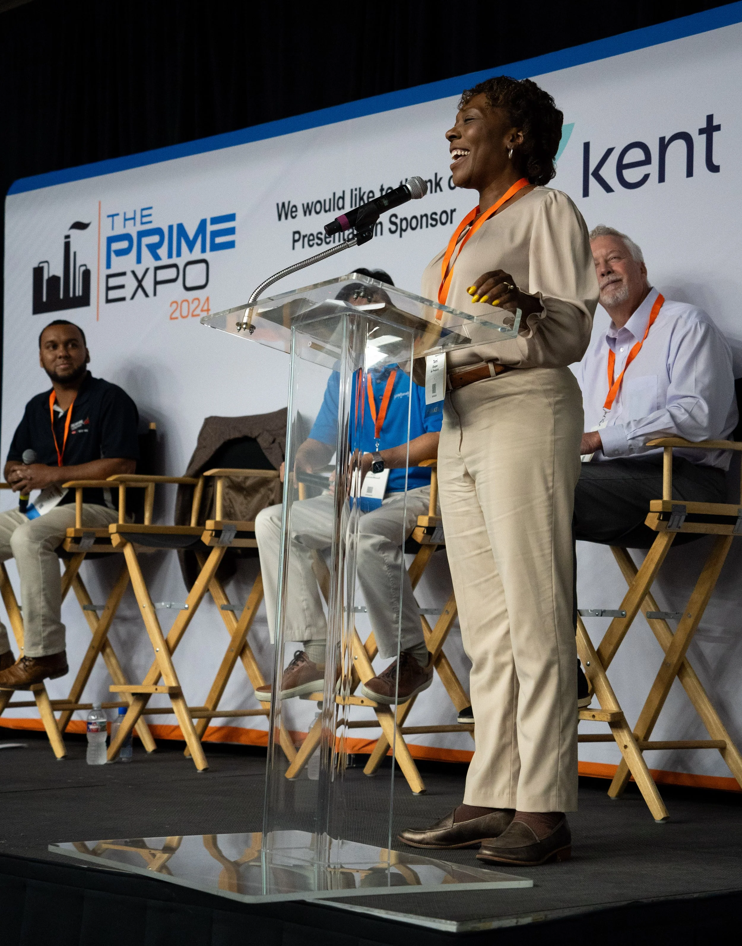 A woman is speaking at a podium during the Prime Expo 2024, with a smiling expression. Three men are seated behind her on the stage, one holding a microphone and all wearing orange lanyards.