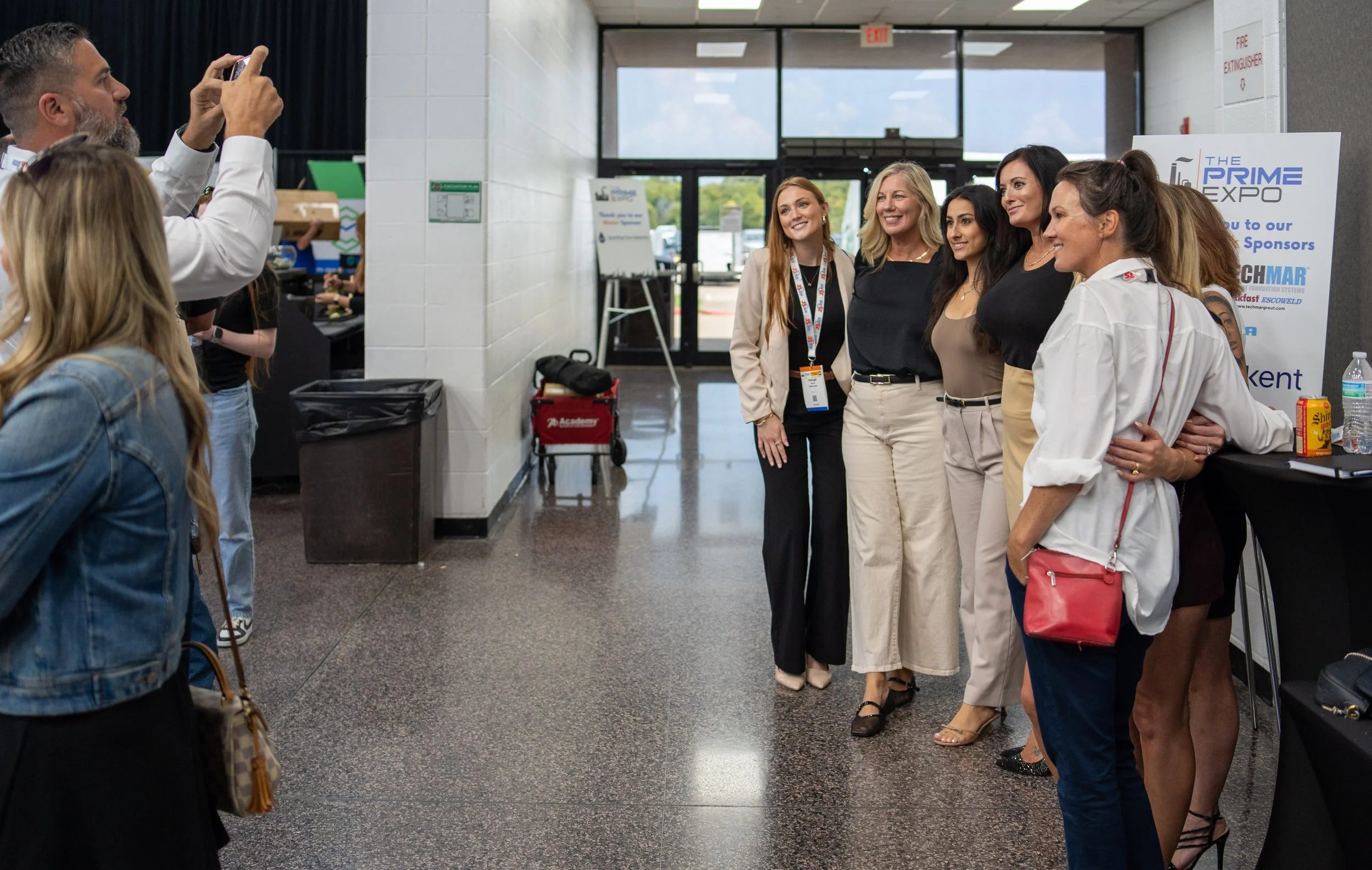 Group of women standing together at a conference or expo, posing for a photo being taken by a man on the left side of the image.