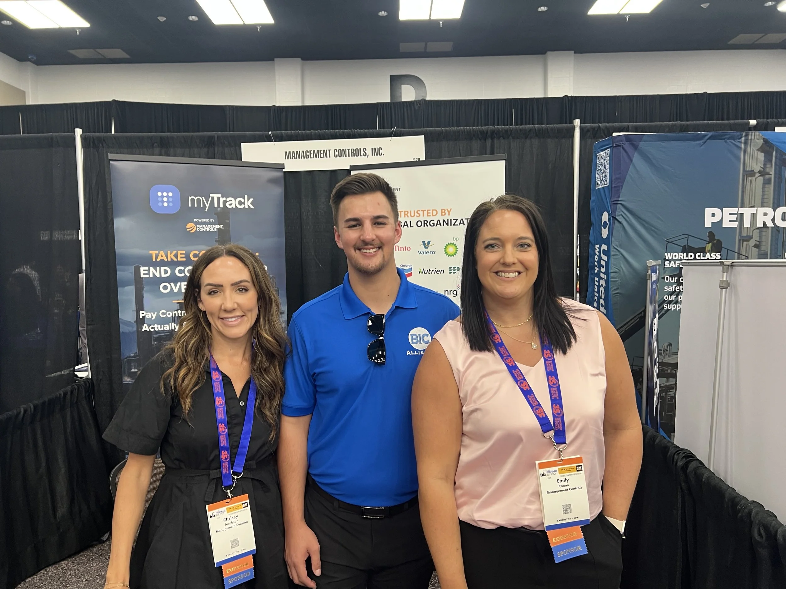 Three people standing together at a trade show or conference, smiling at camera. The woman on the left has long wavy hair, wearing a black dress and a name badge. The man in the middle has short hair, wearing a blue polo shirt with sunglasses hanging