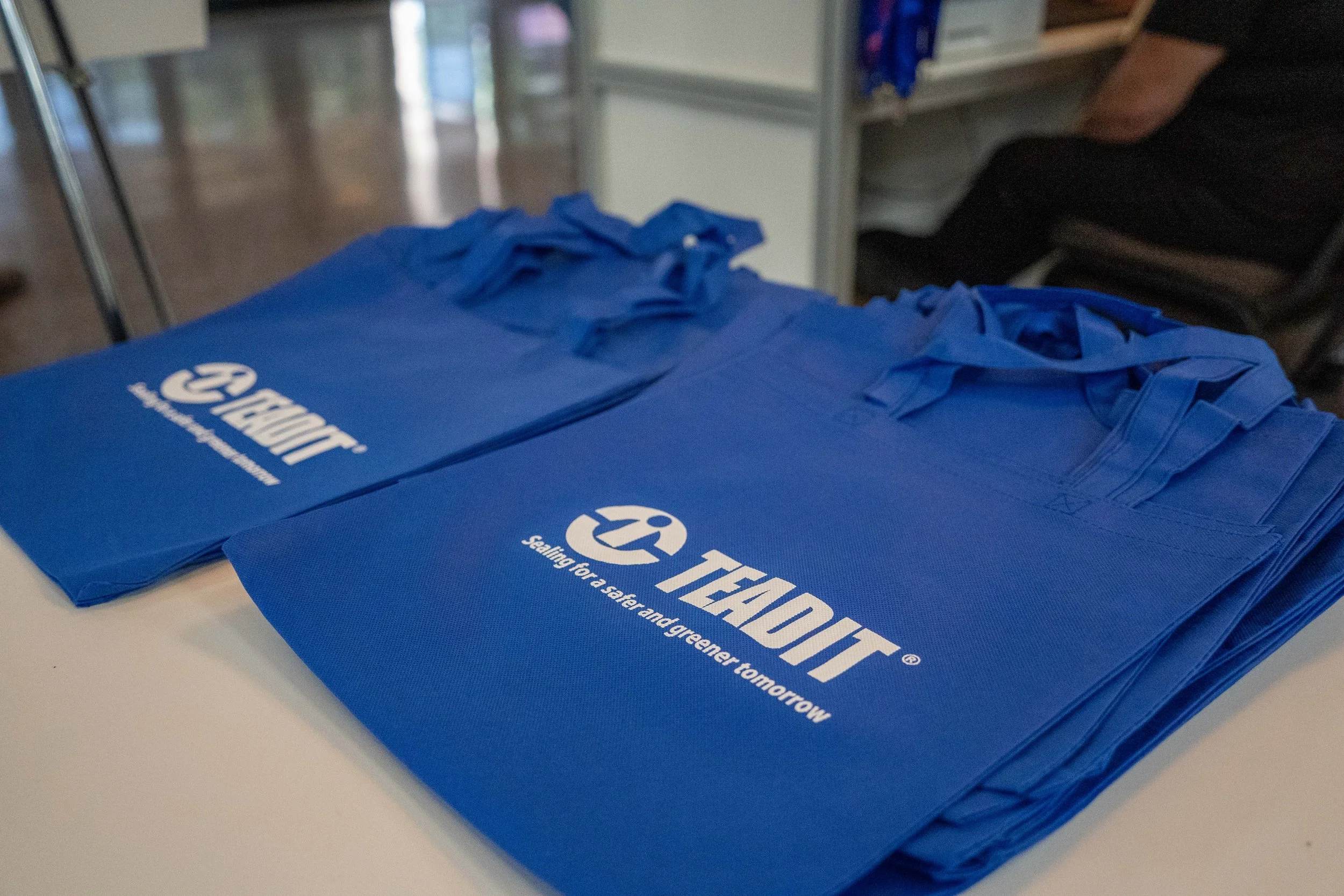 Stack of blue tote bags with the TADDA logo and the slogan 'Sealing for a safer and greener tomorrow' printed on them, placed on a white table.