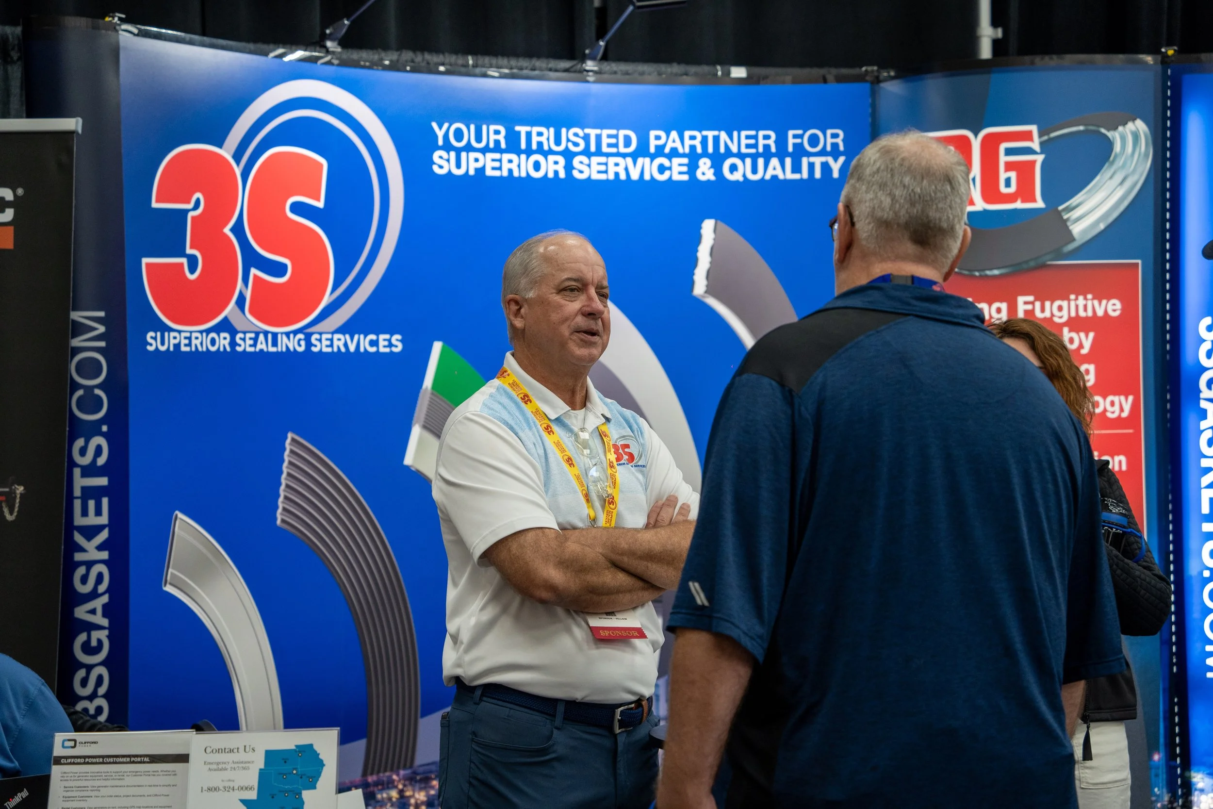Two men having a conversation at a trade show booth for 3S Superior Sealing Services. The background features a large blue banner with the 3S logo and text promoting superior sealing services.