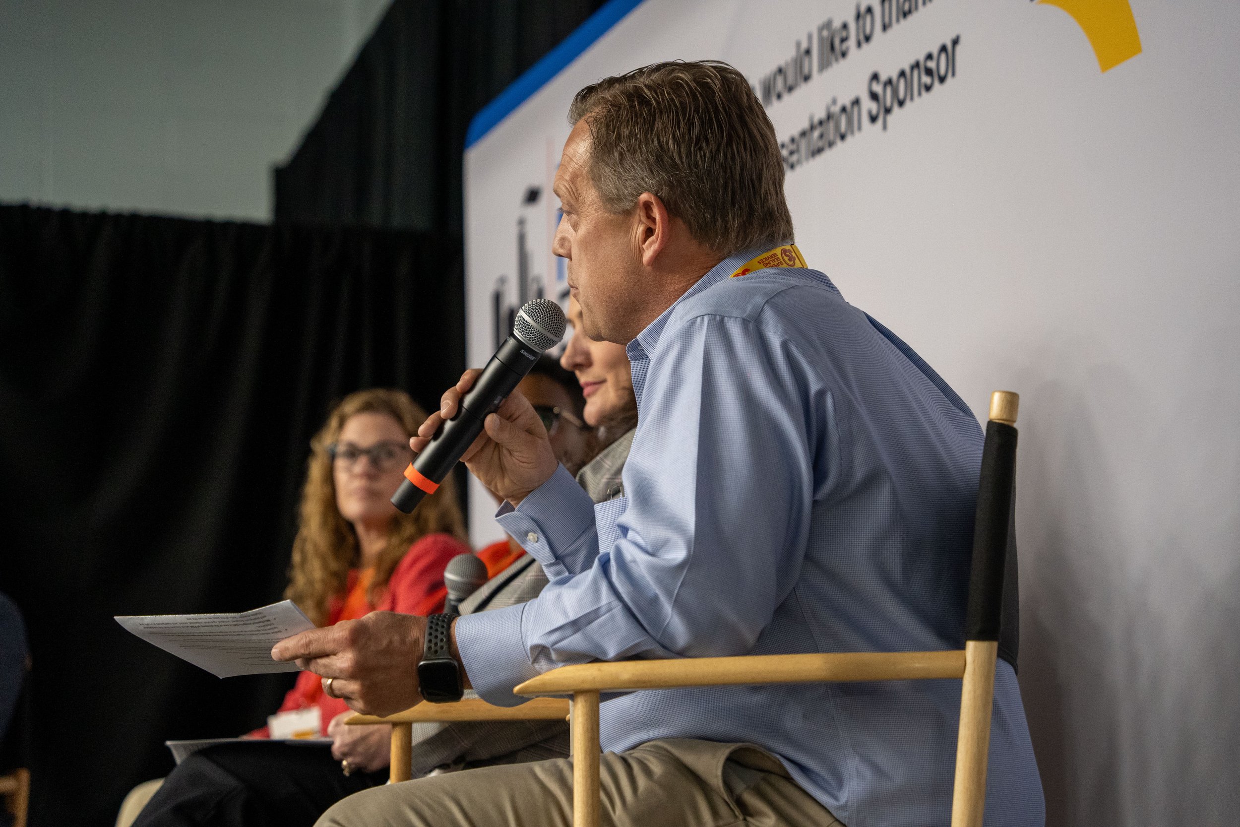 A man holding a microphone and reading from a paper while seated on stage at a panel discussion. Two women are seated beside him, one with curly red hair and glasses, and the other partially visible. The background features a large screen with text a