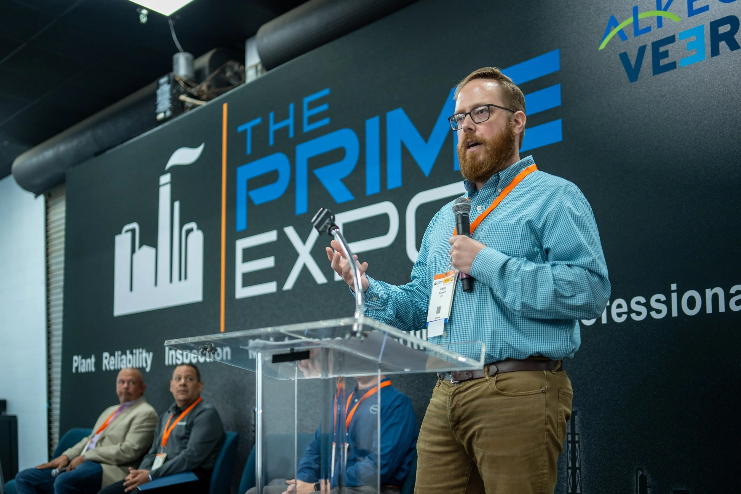 A man with glasses, a beard, wearing a blue checkered shirt and khaki pants, speaking at a conference with a microphone in his right hand and a microphone stand in his left. Behind him is a large banner reading 'The PRIME Expo' with additional words 