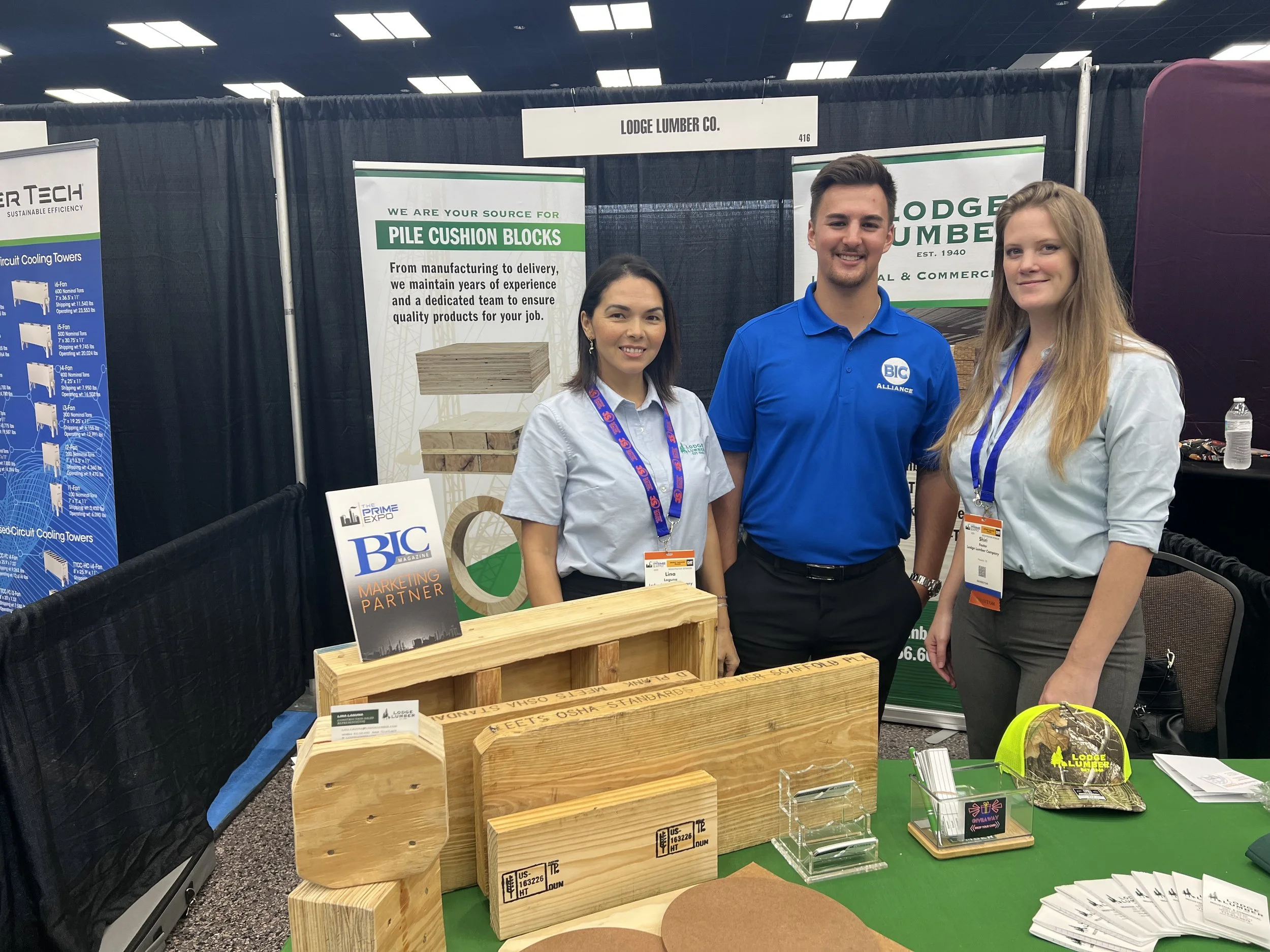 Three people standing behind a booth at a trade show or expo, with banners and wooden products on the table in front of them.
