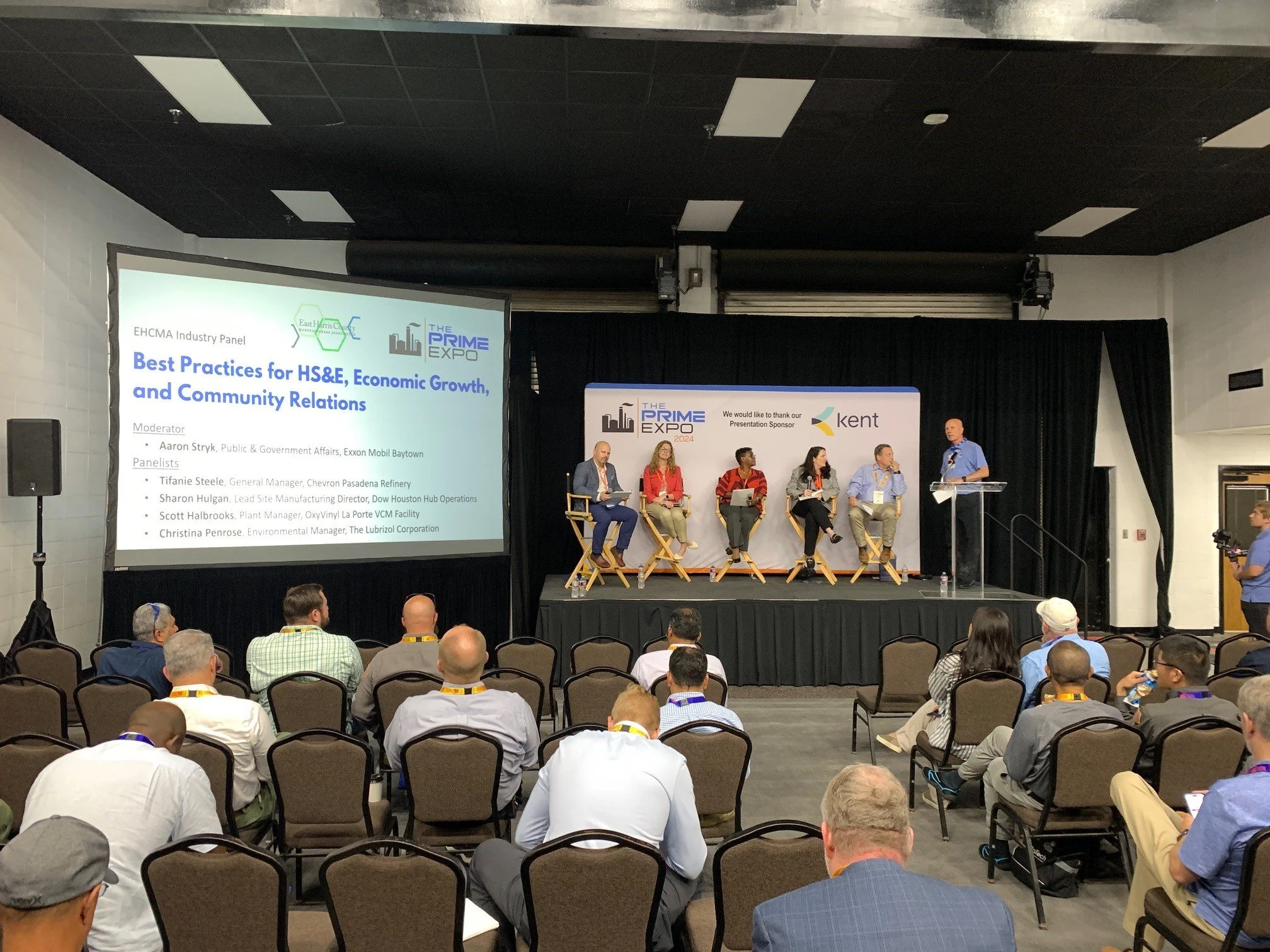 A panel discussion at a conference with five speakers on stage, five seats, and an audience seated facing the stage. The screen displays a presentation titled 'Best Practices for HS&E, Economic Growth, and Community Relations' with moderator and pane