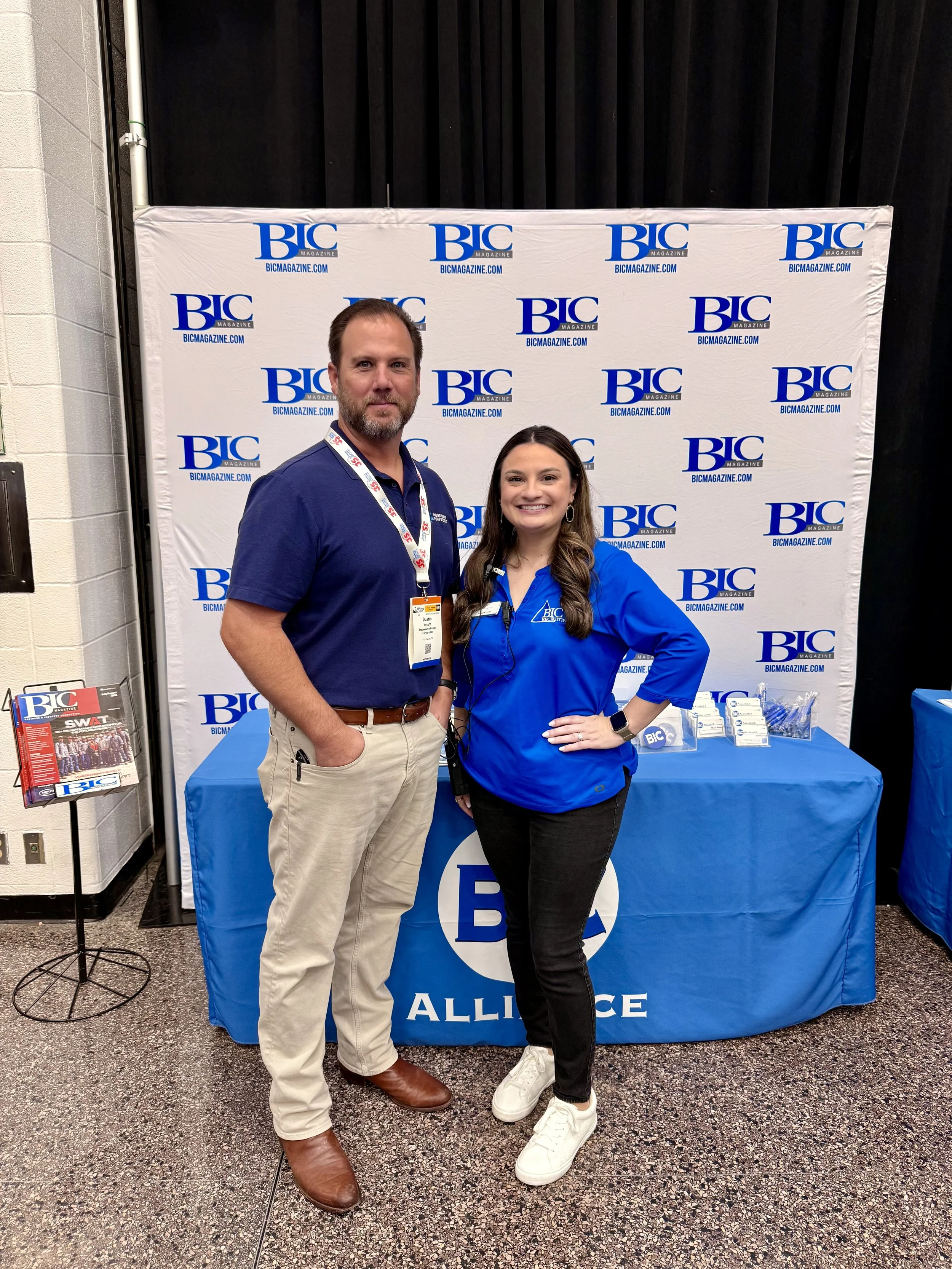 A man and woman standing together in front of a BIC Magazine backdrop at an event. The man has a beard, is wearing a navy blue polo shirt, beige pants, and brown shoes. The woman has long dark hair, is wearing a blue BIC jacket, black pants, white sn