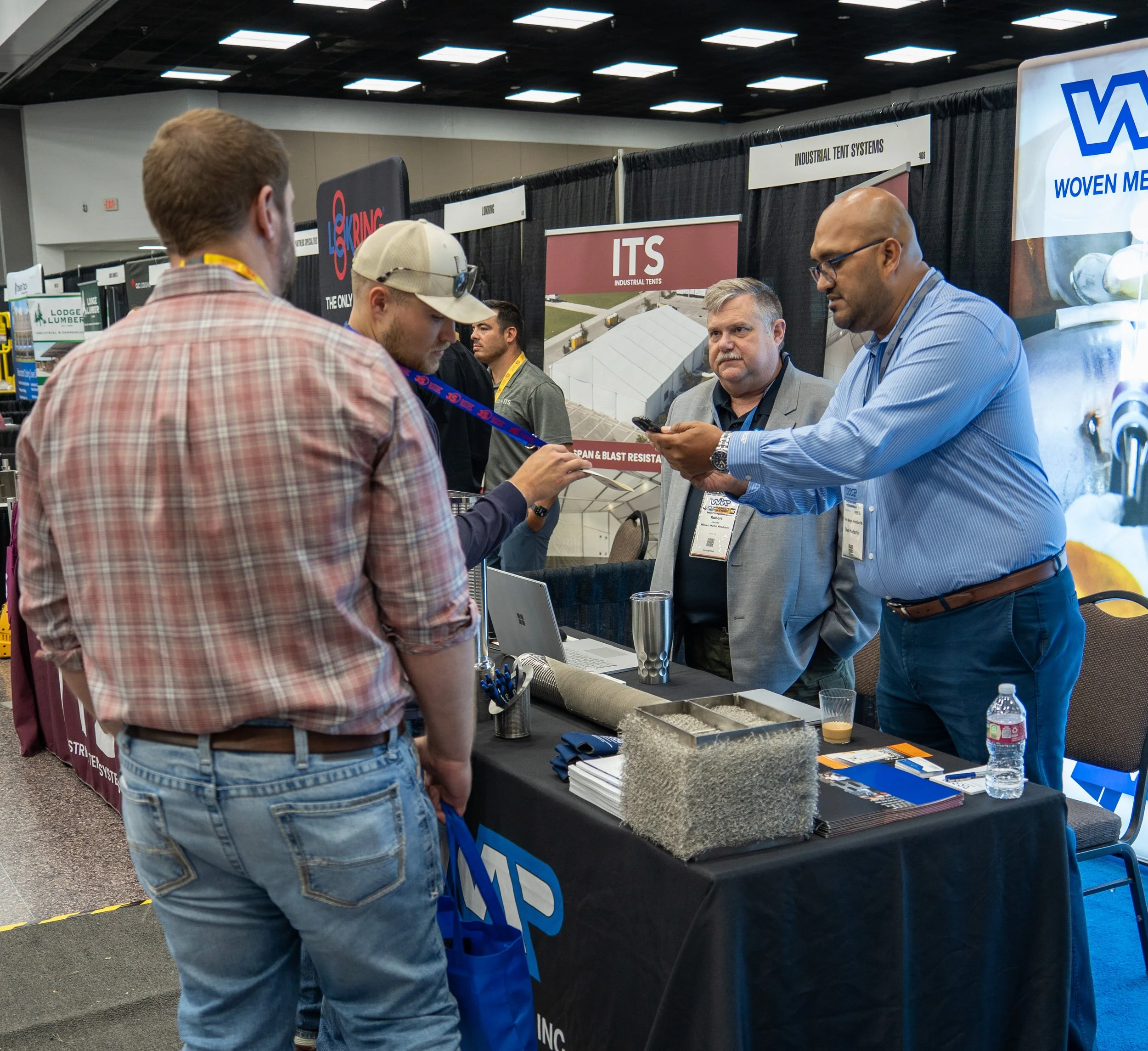 A trade show booth with three men engaged in conversation, one man is showing a product on his phone, other two men are looking attentively. The booth has promotional banners and a table with brochures, a laptop, and samples.