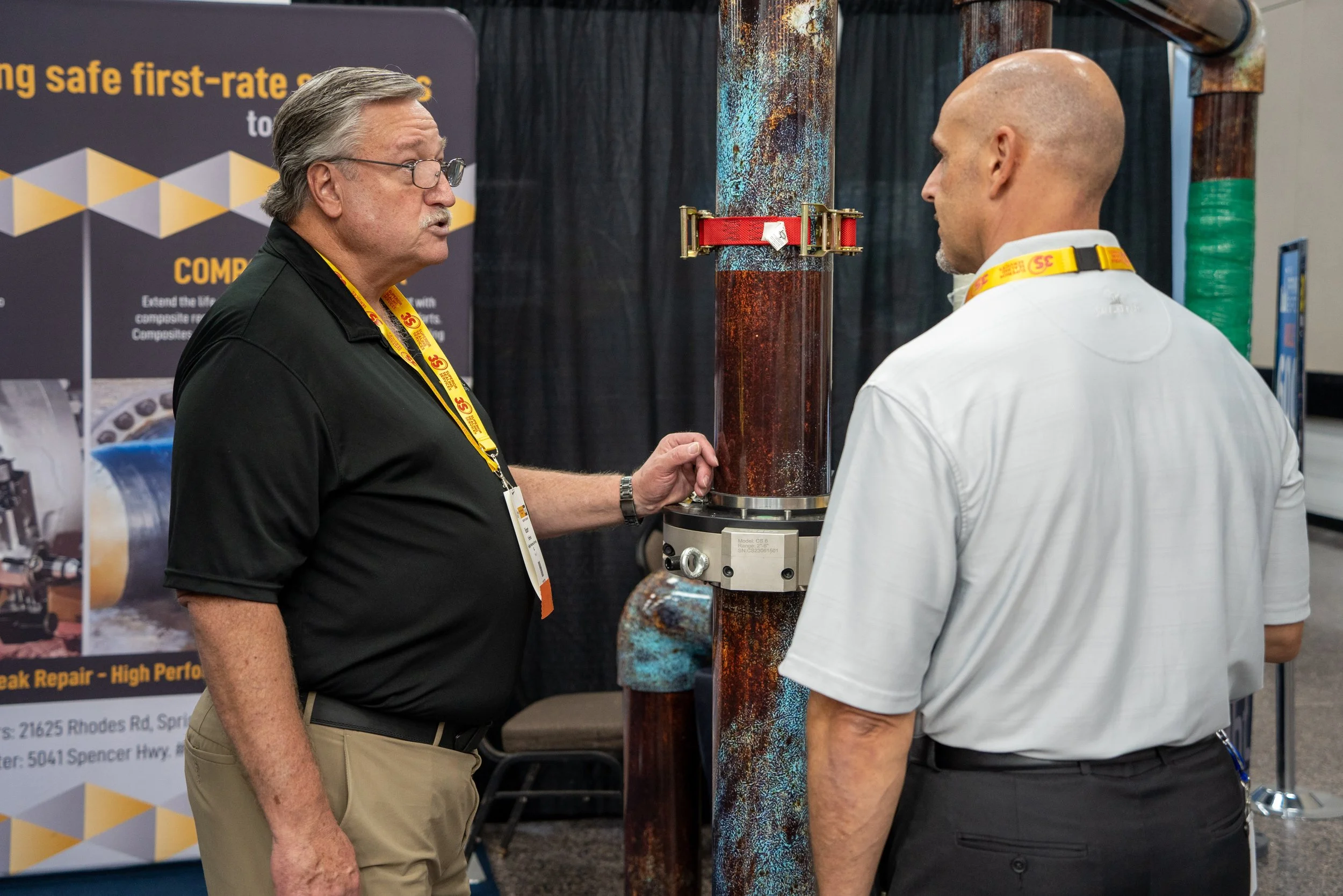 Two men are examining a large rusted industrial pipe with a measuring device attached, standing in front of a black display banner at an exhibition.