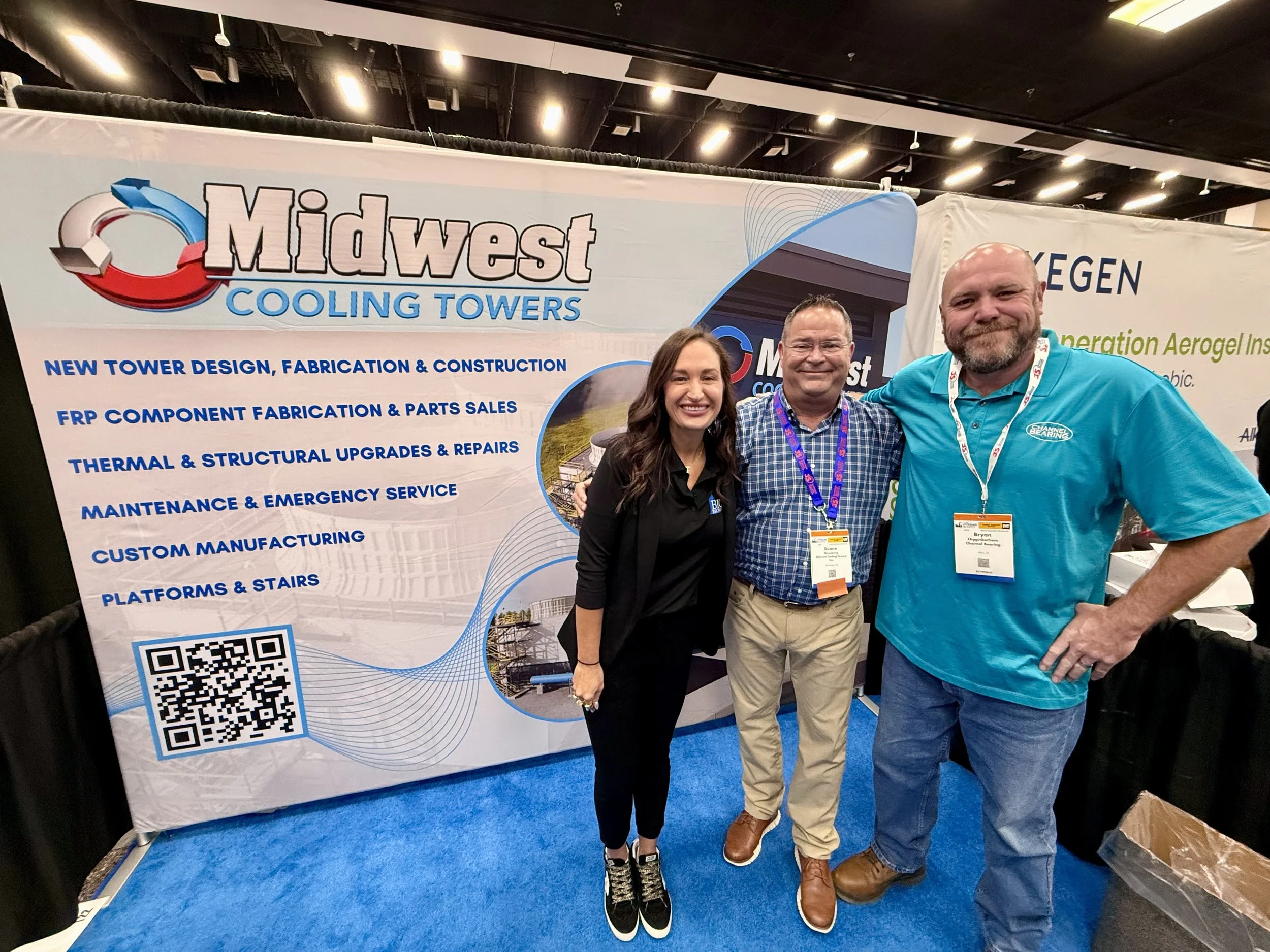 Three people standing in front of a booth at a trade show, smiling at the camera. The booth banner displays the logo and services of Midwest Cooling Towers, including new tower design, fabrication, repairs, and custom manufacturing.