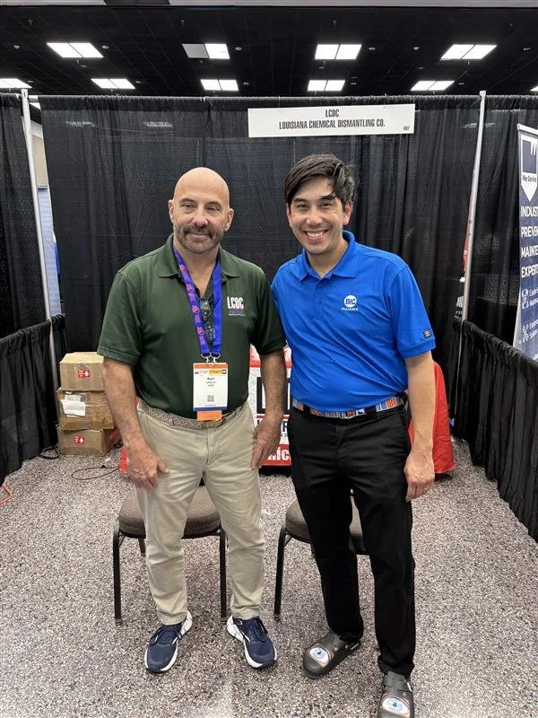 Two men standing in front of a black booth at an industry event, smiling at the camera. One is wearing a green polo shirt with a badge and lanyard, the other is in a blue polo shirt. Behind them is a sign for Louisiana Chemical Dismantling Co.