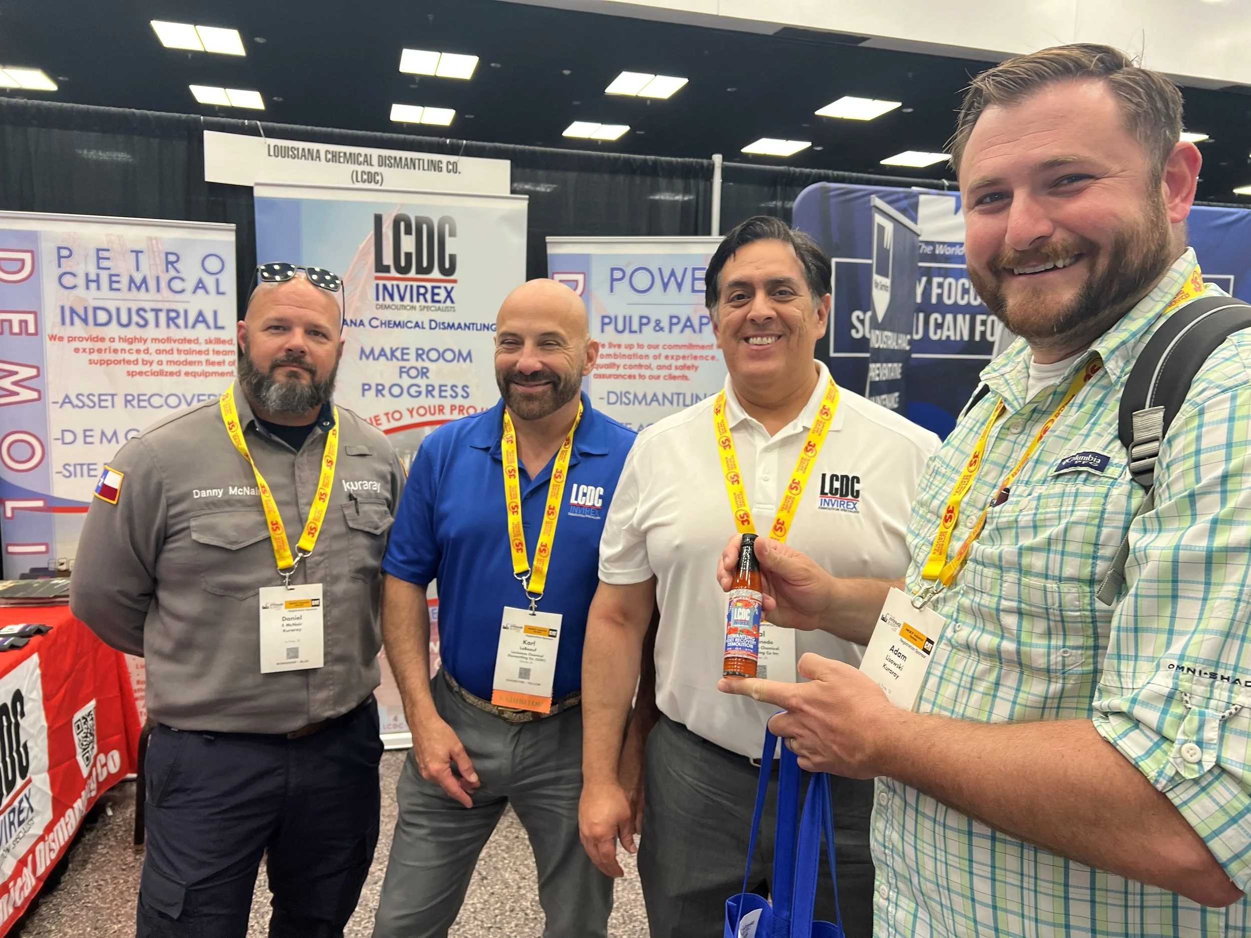Four men at a trade show booth smiling, with banners behind them displaying company names like Louisiana Chemical Dismantling Co. and Petro Chemical Industrial, some wearing yellow lanyards with event passes.